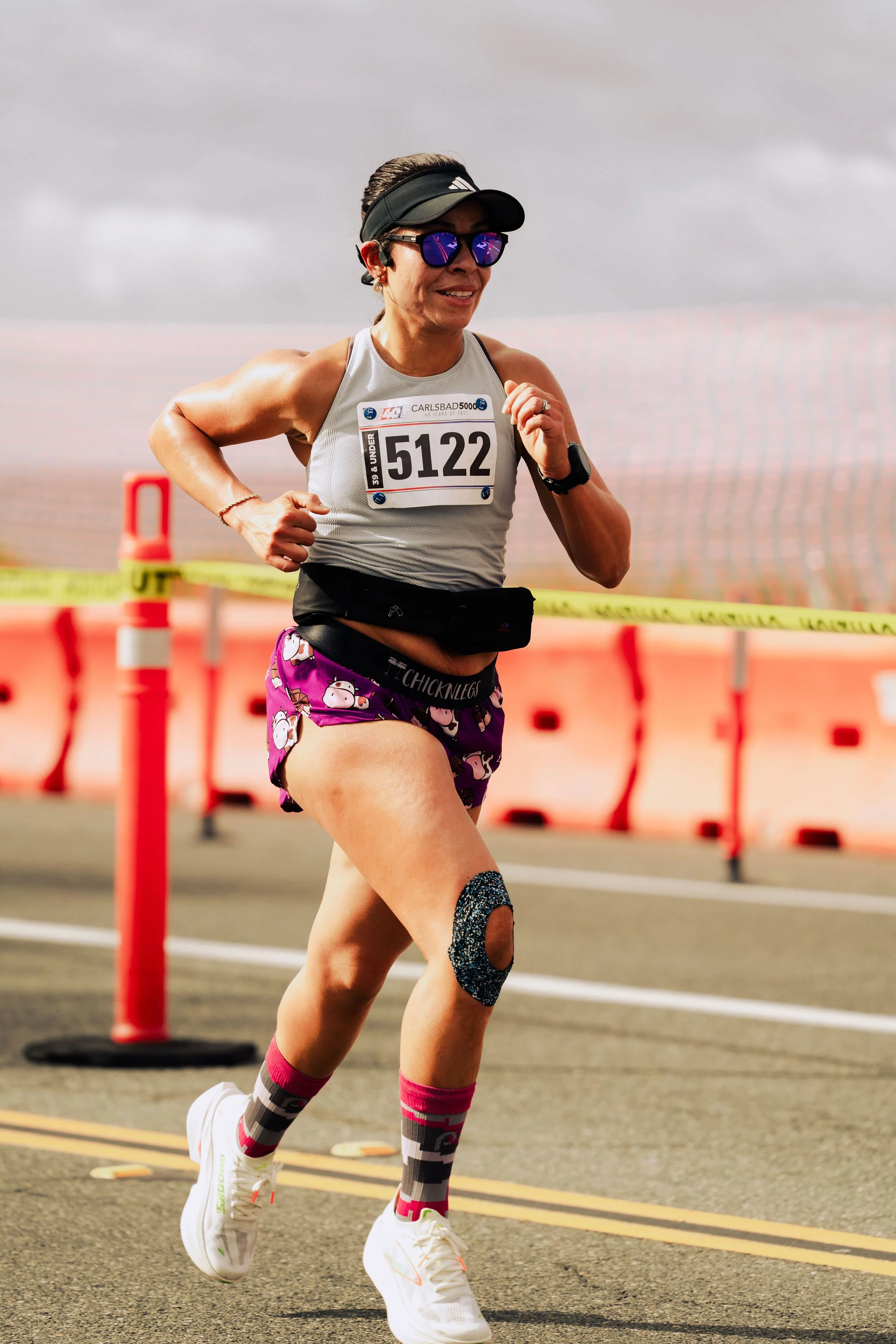 A woman marathon runner running on a city street, wearing sunglasses, a watch, a gray tank top with race number 5122, pink and purple shorts with a cow pattern, white running shoes, and knee-high socks with gray, pink, and black stripes. There are or