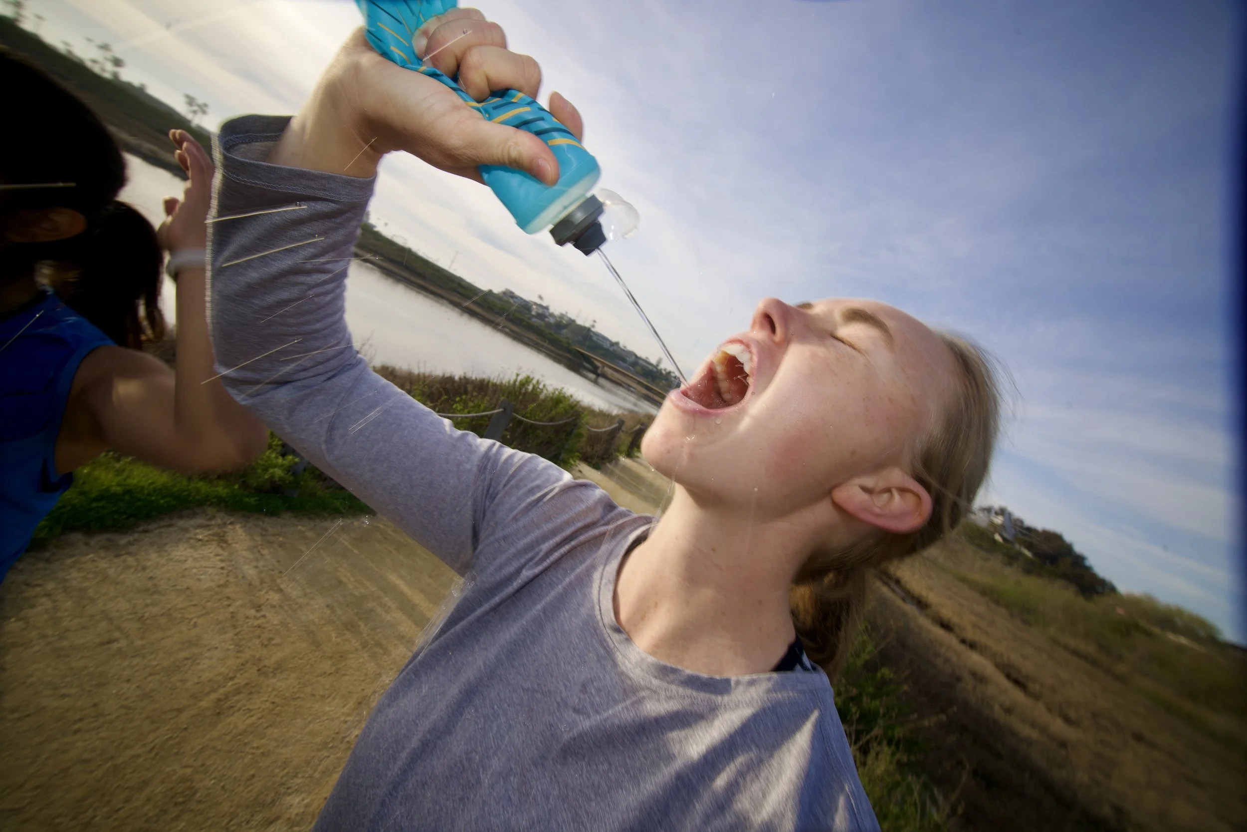 A young girl with red hair, wearing a gray shirt, holding a water bottle with a drop of water coming out, outdoors during daytime.