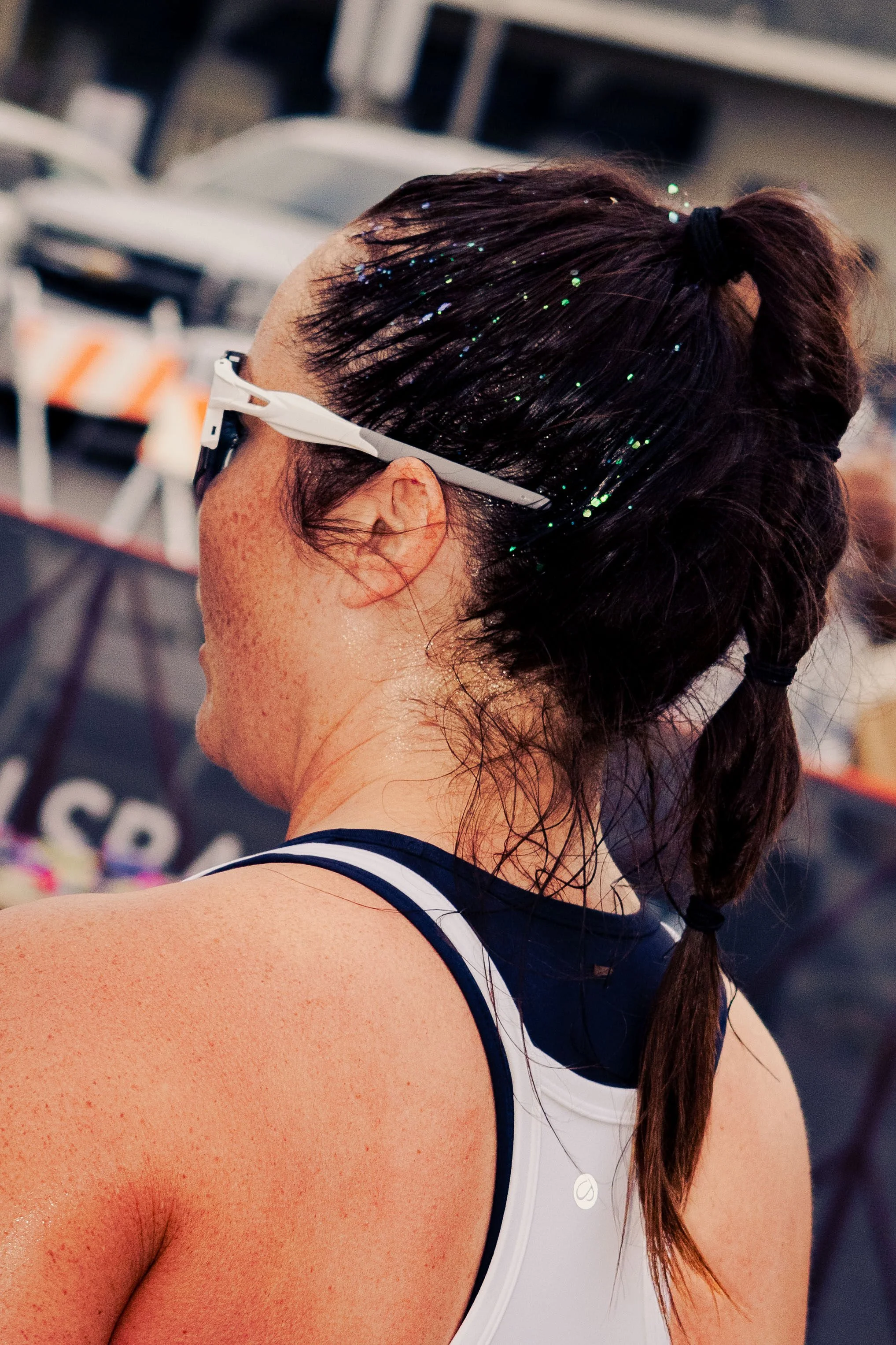 Close-up of a woman with brown hair tied in a ponytail, wearing white sports glasses and a white athletic top, outdoor setting with blurred background.