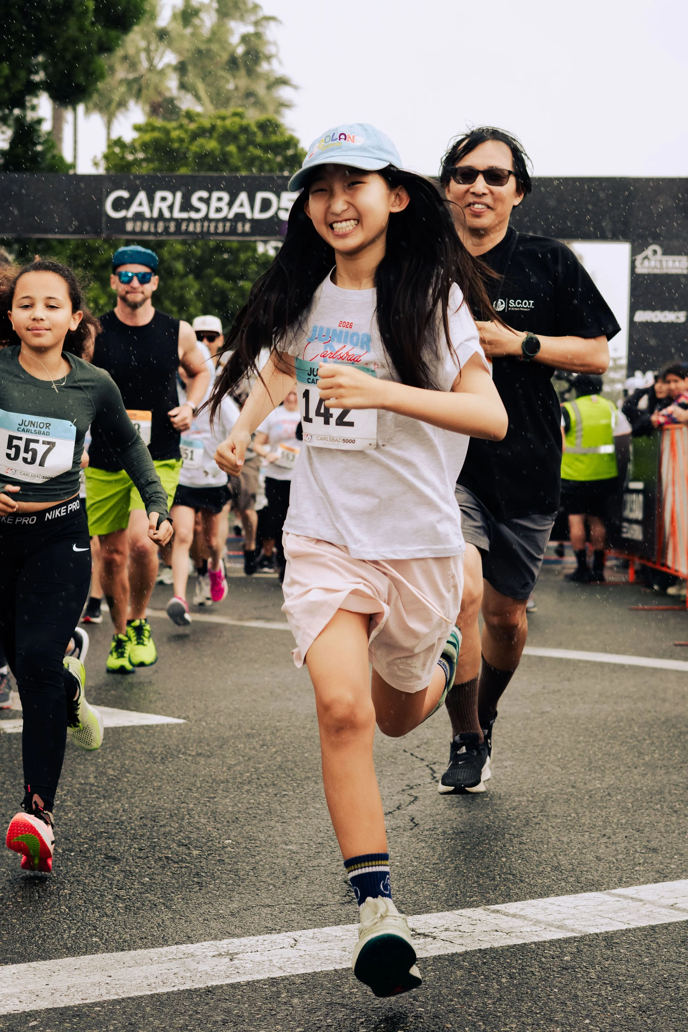Young girl running in a race, smiling, wearing a white shirt, pink shorts, a blue cap, and sports shoes. Several other runners and a finish line are visible in the background.