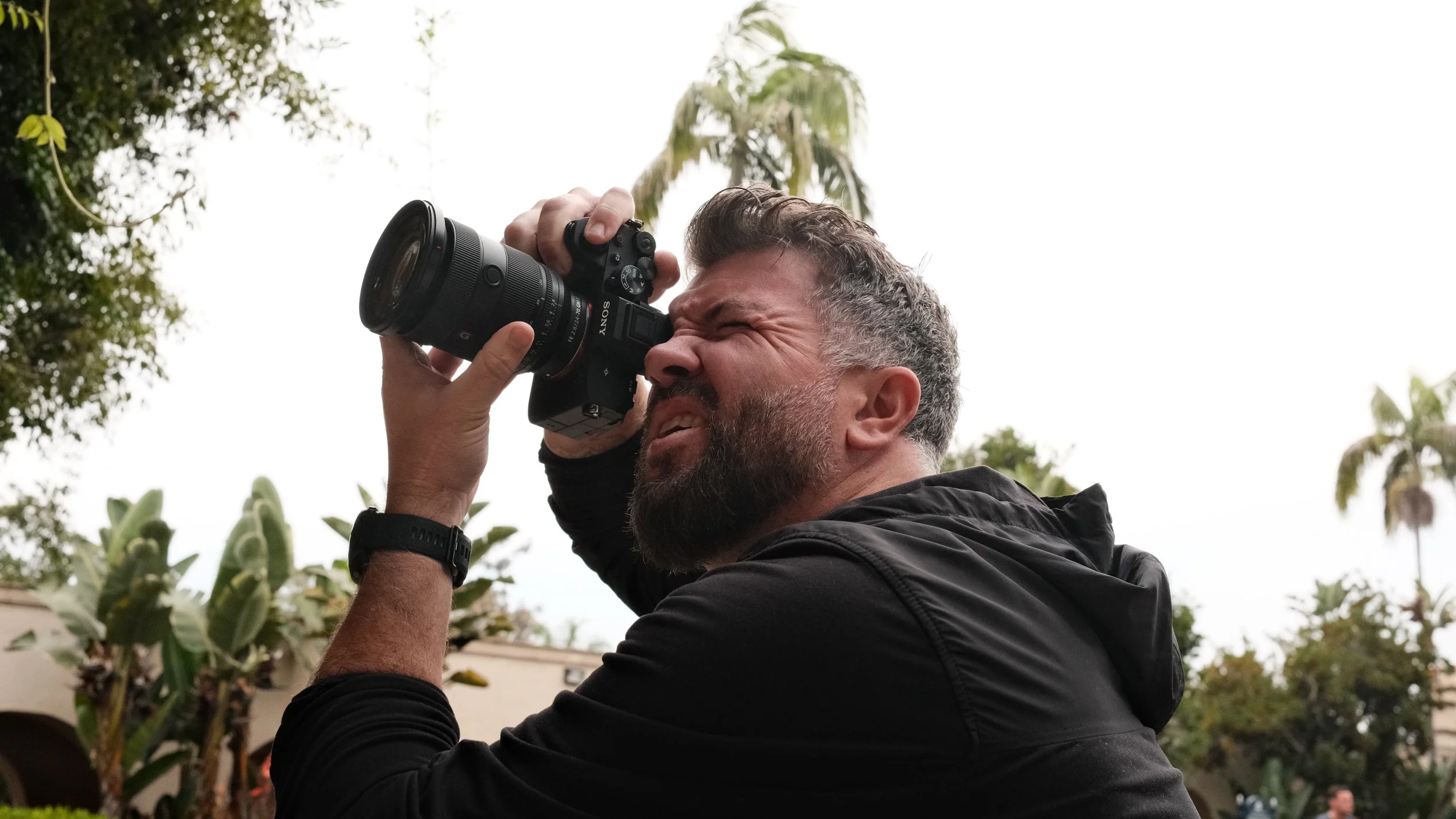 A man with short gray hair and a beard is holding a Sony camera up to his eye, taking a photo outdoors. He is wearing a black jacket, and the background shows trees and plants with an overcast sky.