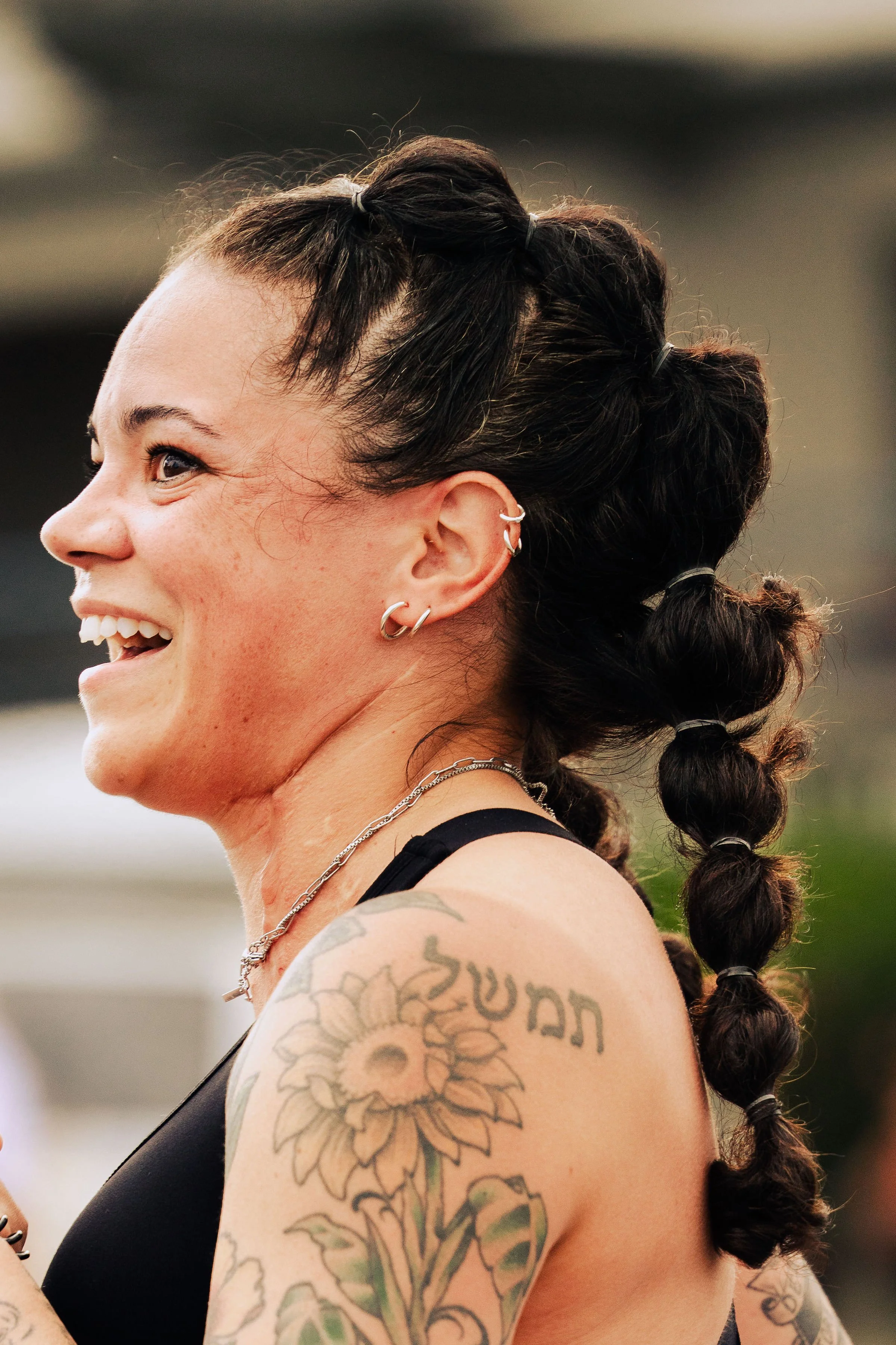 A woman with dark hair styled in bun pigtails, smiling, wearing multiple earrings, a silver necklace, and a black sleeveless top, showing tattoos on her upper arm and shoulder.