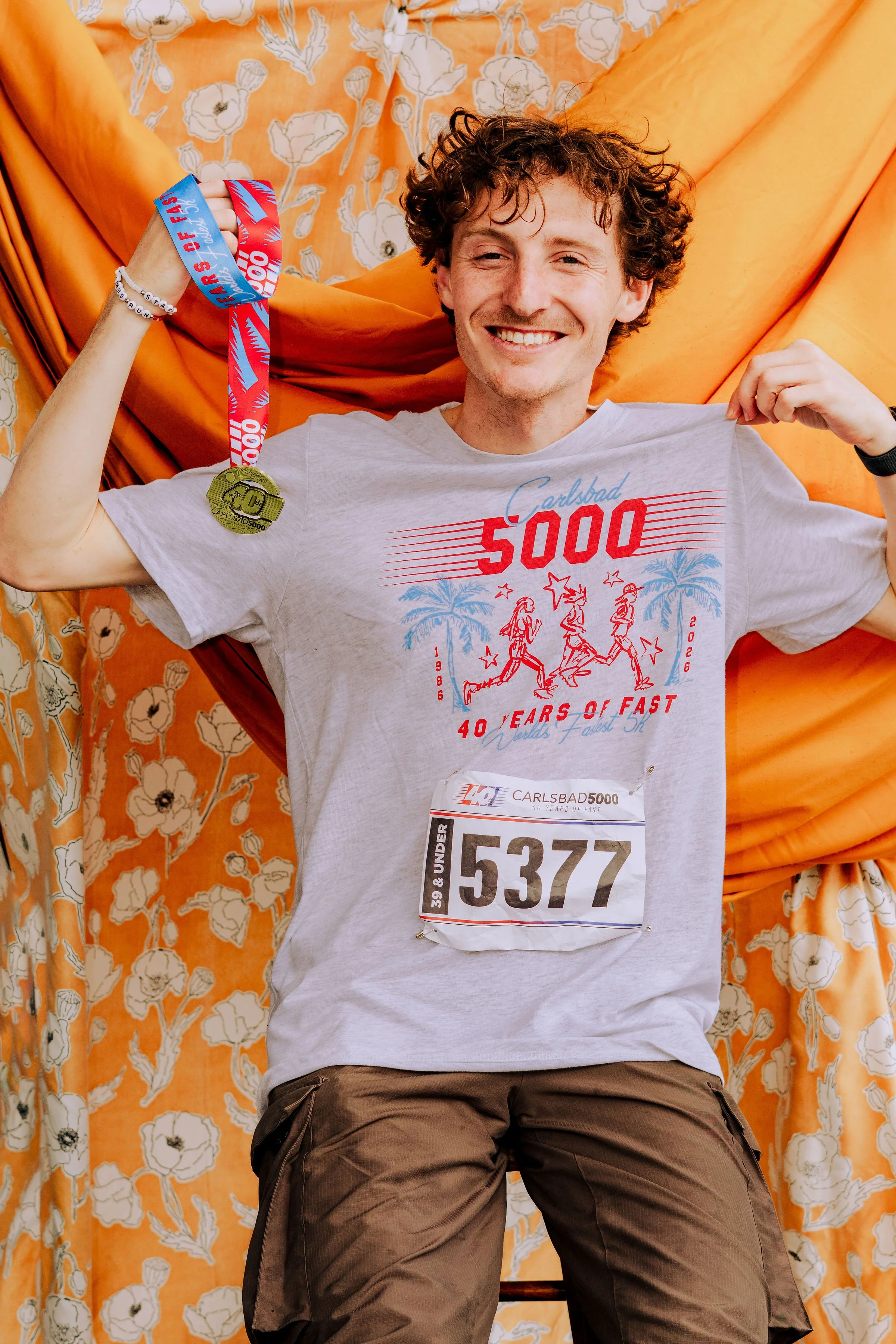Man lying on a bed with orange floral bedding, smiling, wearing a white t-shirt with red and blue print, holding up a finisher medal.