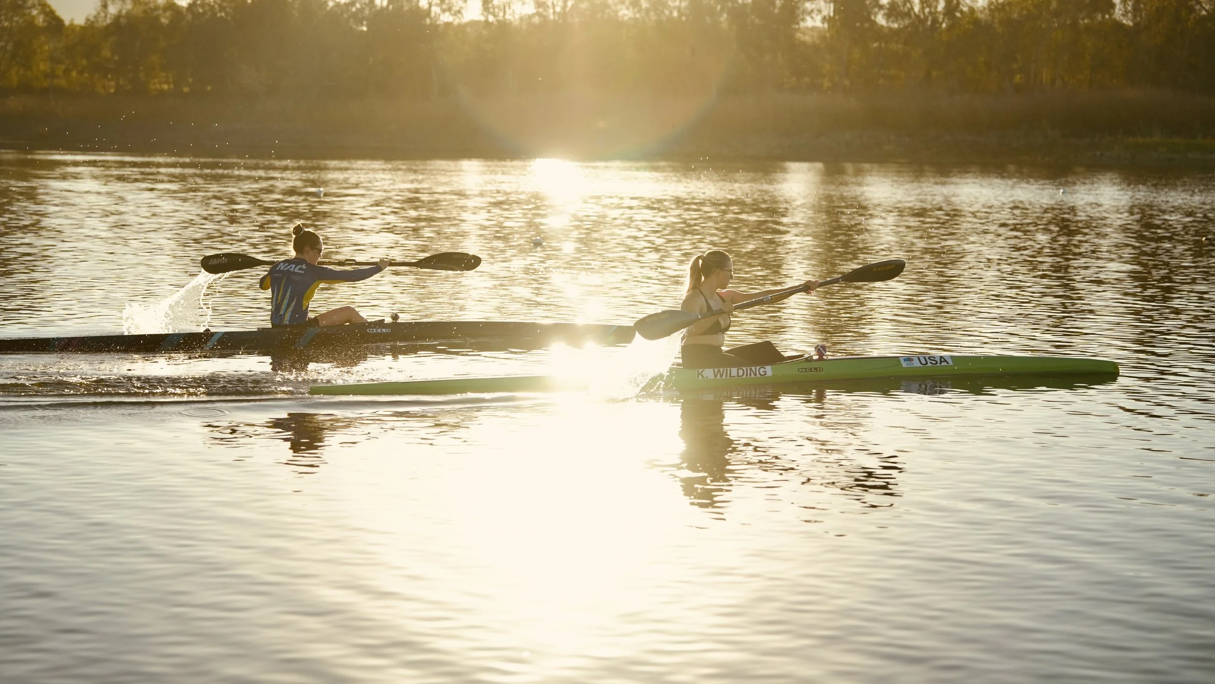 Two women kayaking on a calm river during sunset, with reflections and trees in the background.