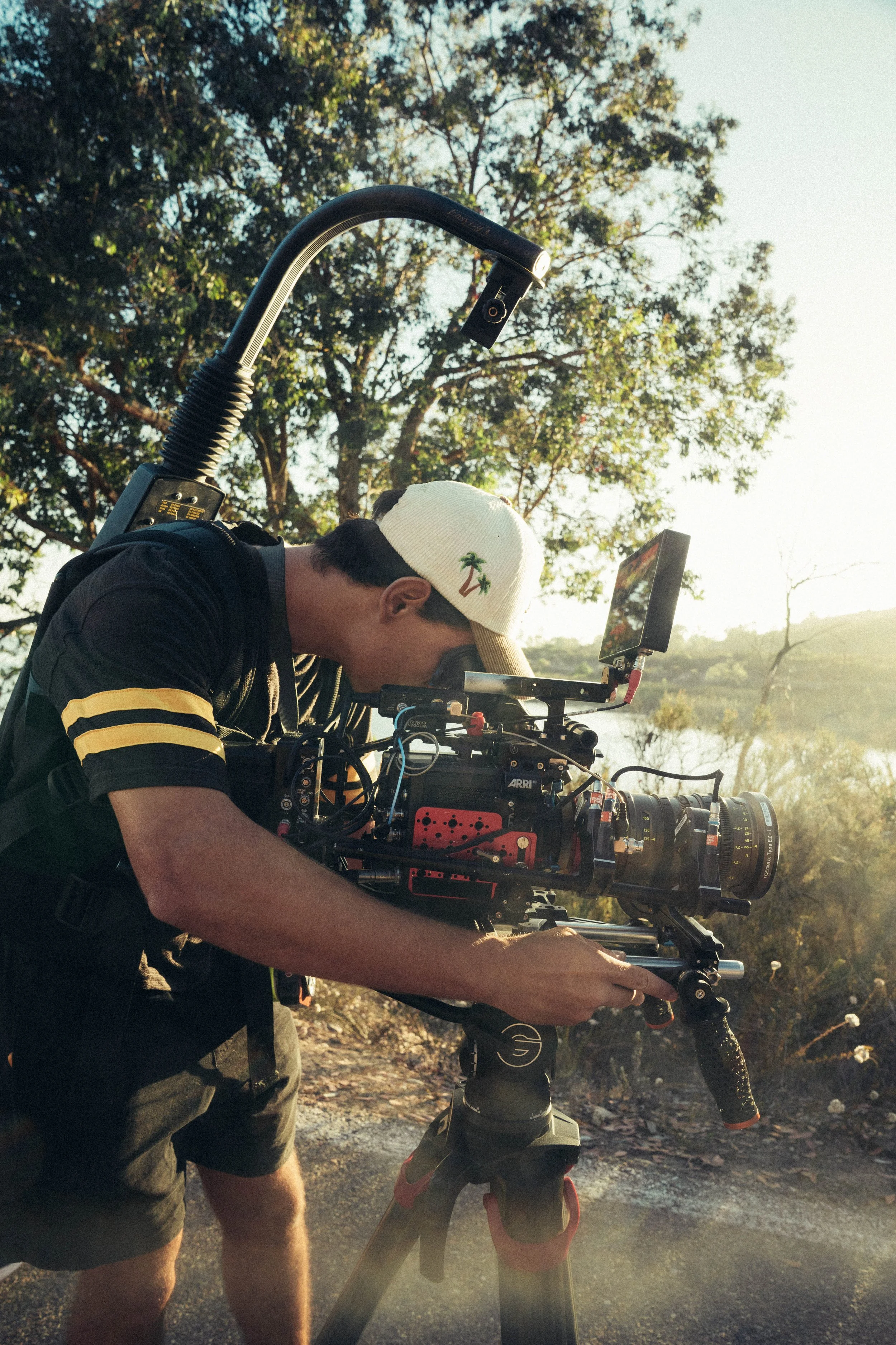 A person wearing a white cap with a palm tree embroidered on it is looking through a camera mounted on a bicycle. The background features trees and a bright sky.