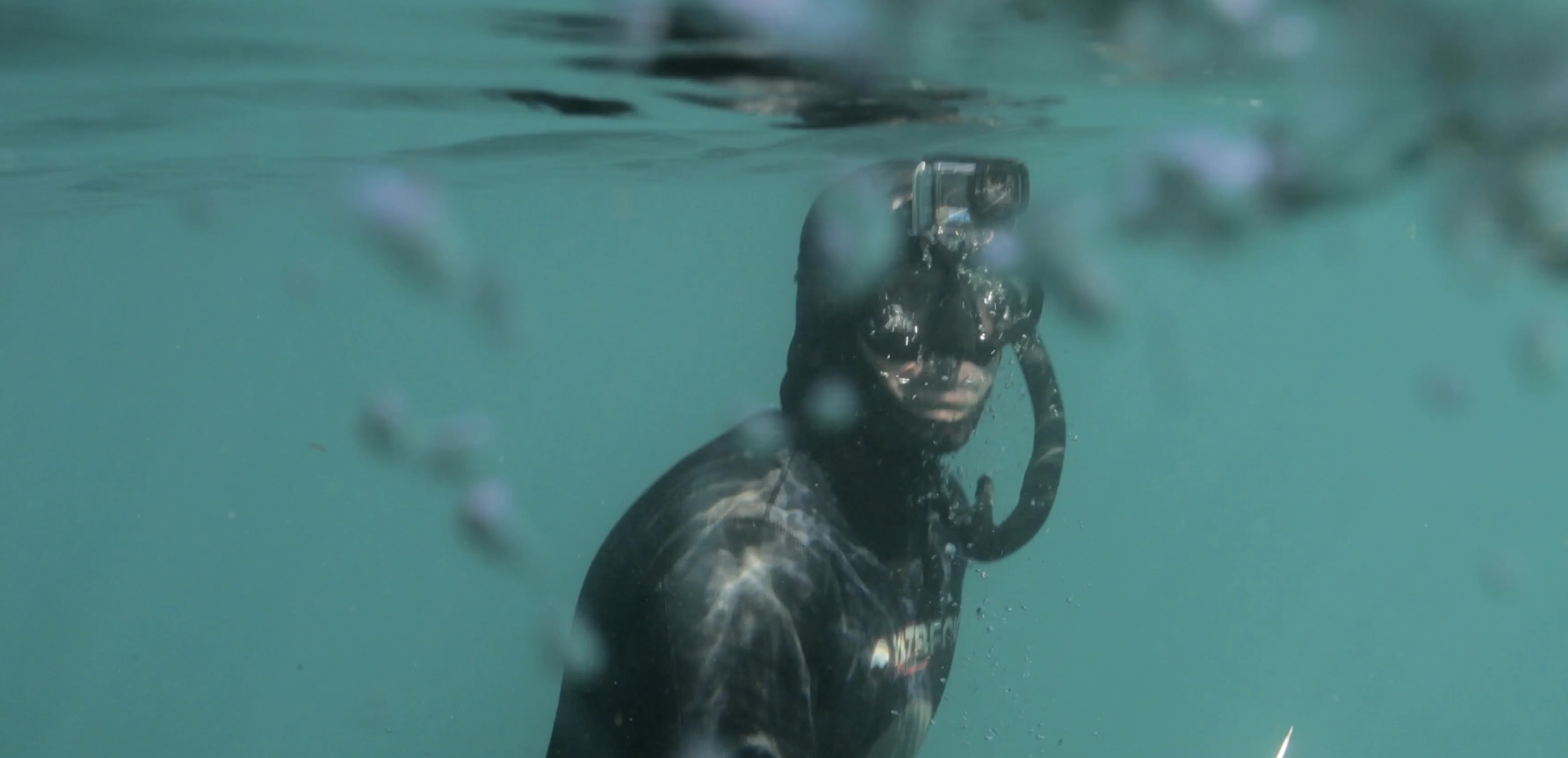 A person in a wetsuit snorkeling underwater with a diving mask and snorkel, looking towards the camera.
