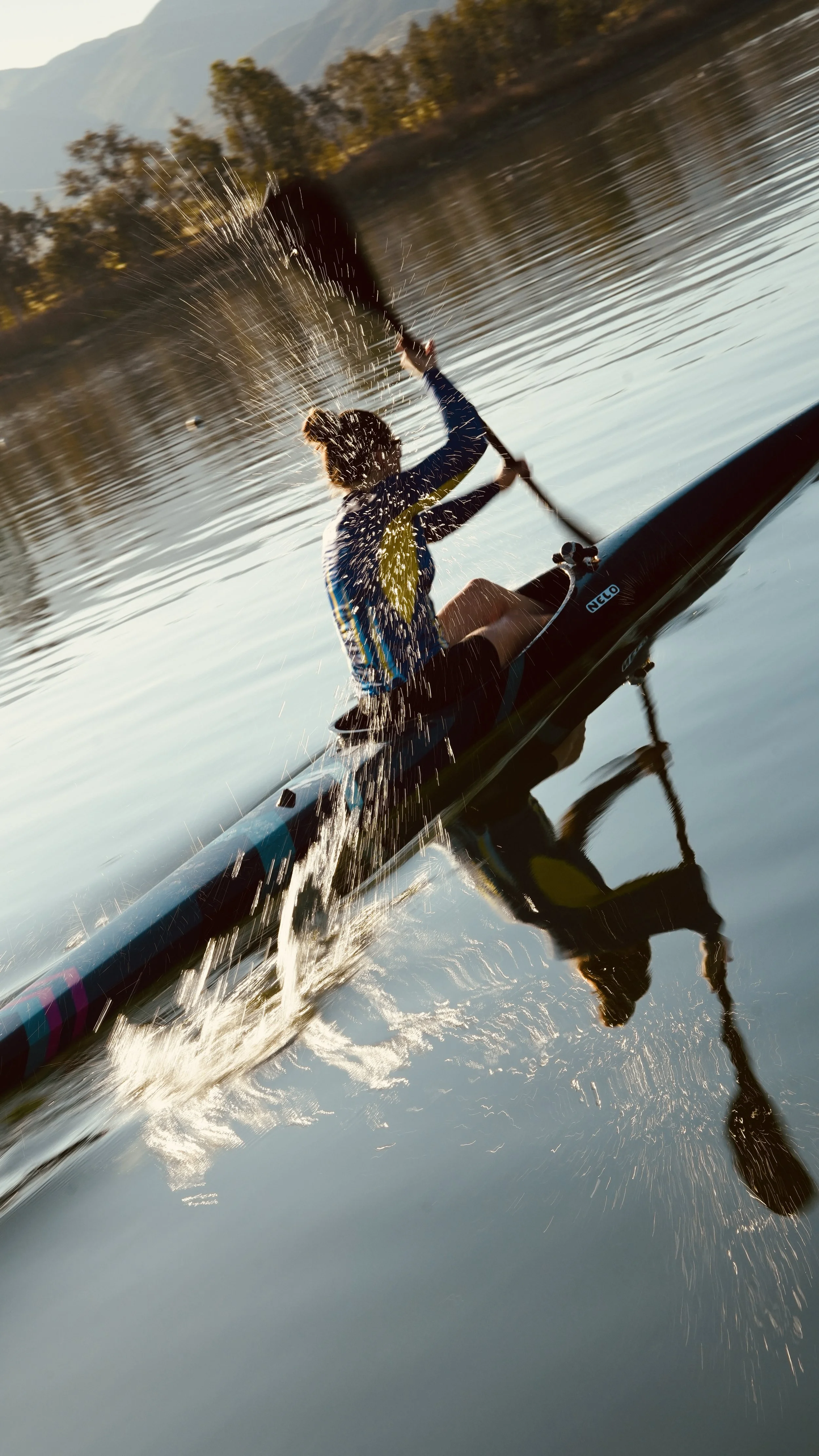 A person kayaking on a calm river during sunset, creating a splash of water as they paddle, with mountains and trees in the background.