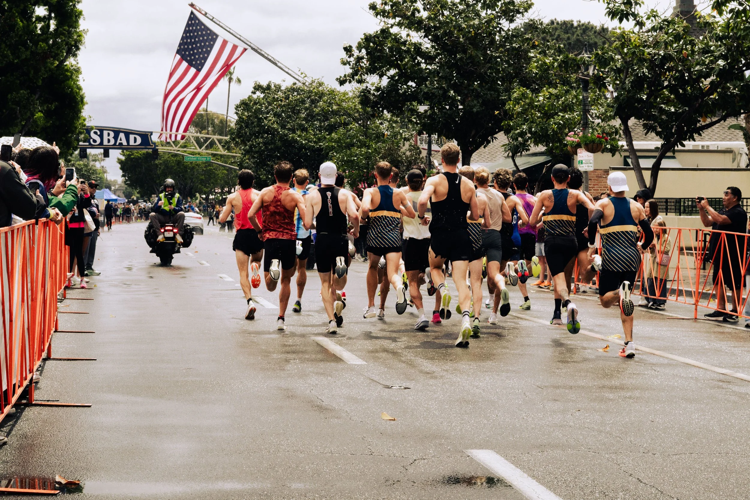 A group of marathon runners participating in a race on a city street, with spectators on the side and an American flag hanging above.