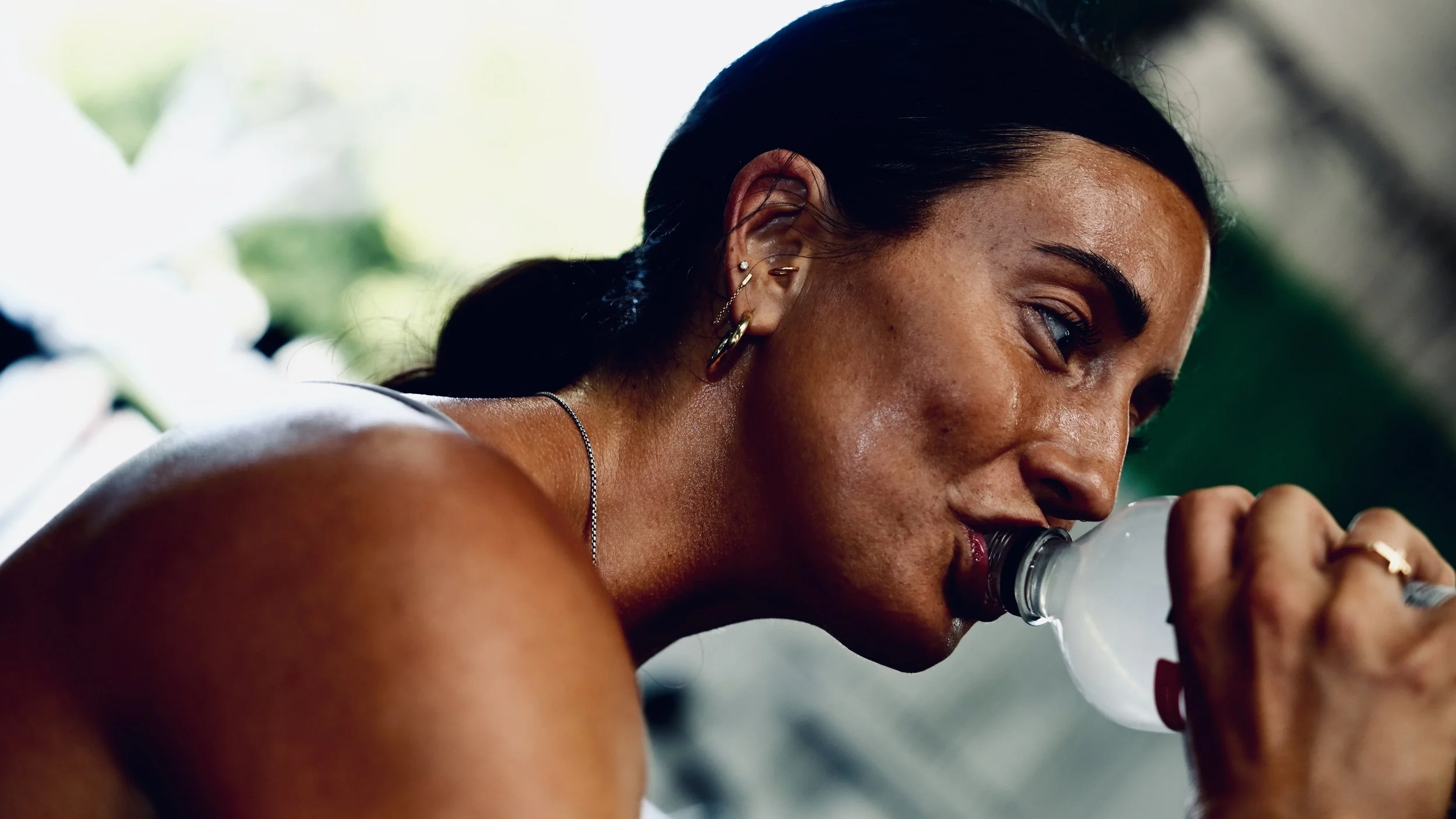Woman with dark hair, multiple earrings, and tattoos, drinking water from a clear bottle outdoors.