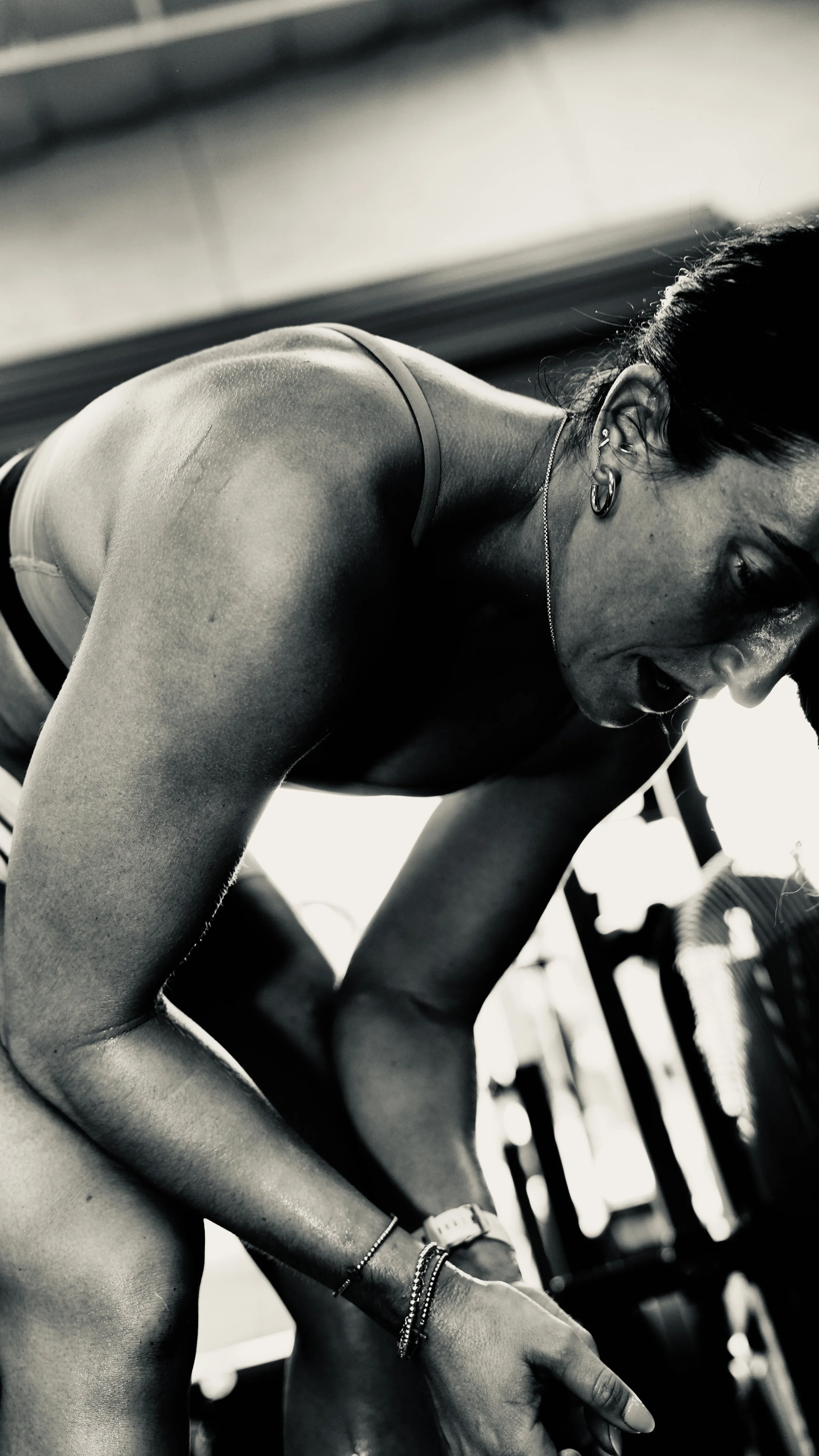 A woman in athletic gear is exercising in a gym, bending forward with her hand on the floor, sweat visible on her skin, wearing earrings and bracelets.