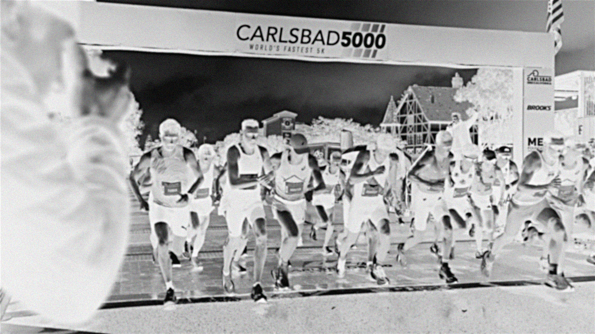 A group of runners participating in a race at the Carlsbad 5000 running event, with a banner overhead and a bridge structure in the background.