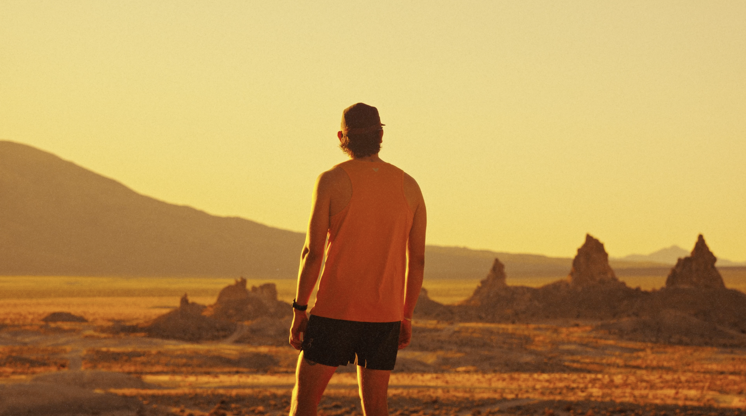 A person in an orange shirt and black shorts standing in a desert landscape during sunset, with rock formations and mountains in the background.