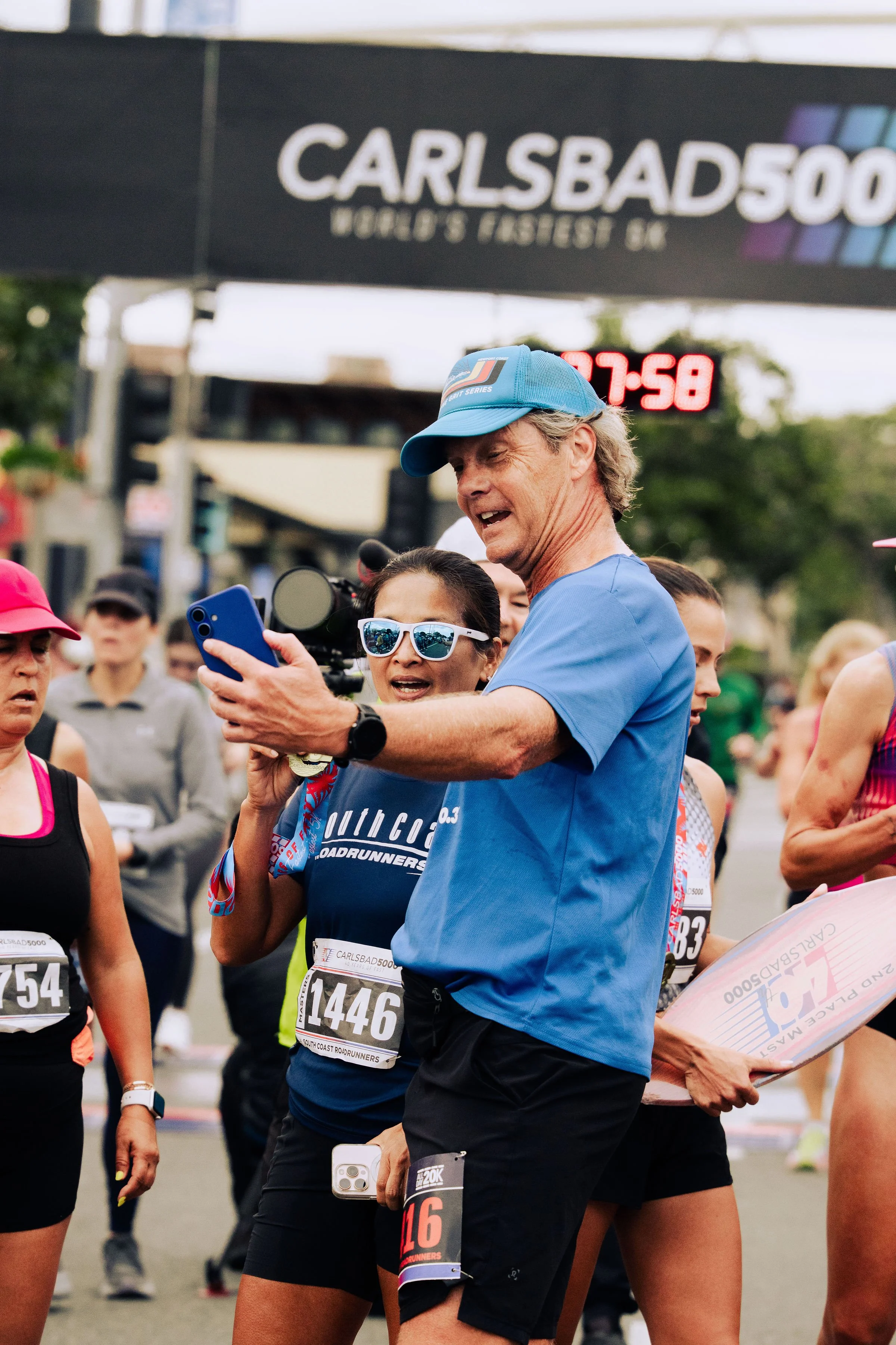 Group of marathon runners gathered at the starting line, wearing race bibs, some with medals, and surrounded by spectators. A man in a blue shirt and cap is looking at his phone. The event is the Carlsbad 5000 running race.