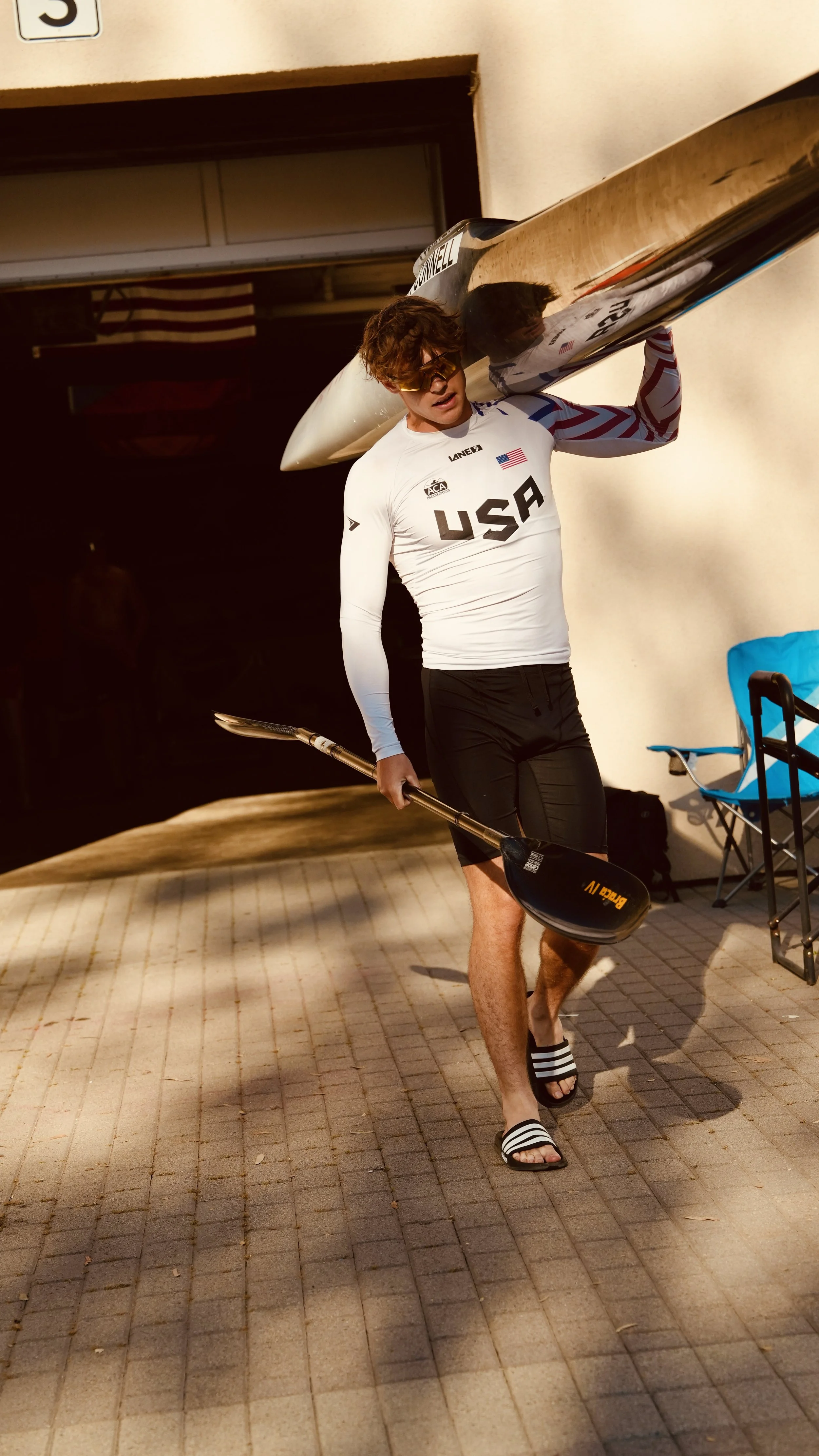 A young man wearing sunglasses, a USA sports shirt, and slide sandals carrying a kayak on his shoulder, holding a paddle in his other hand, walking on a paved surface during daytime.