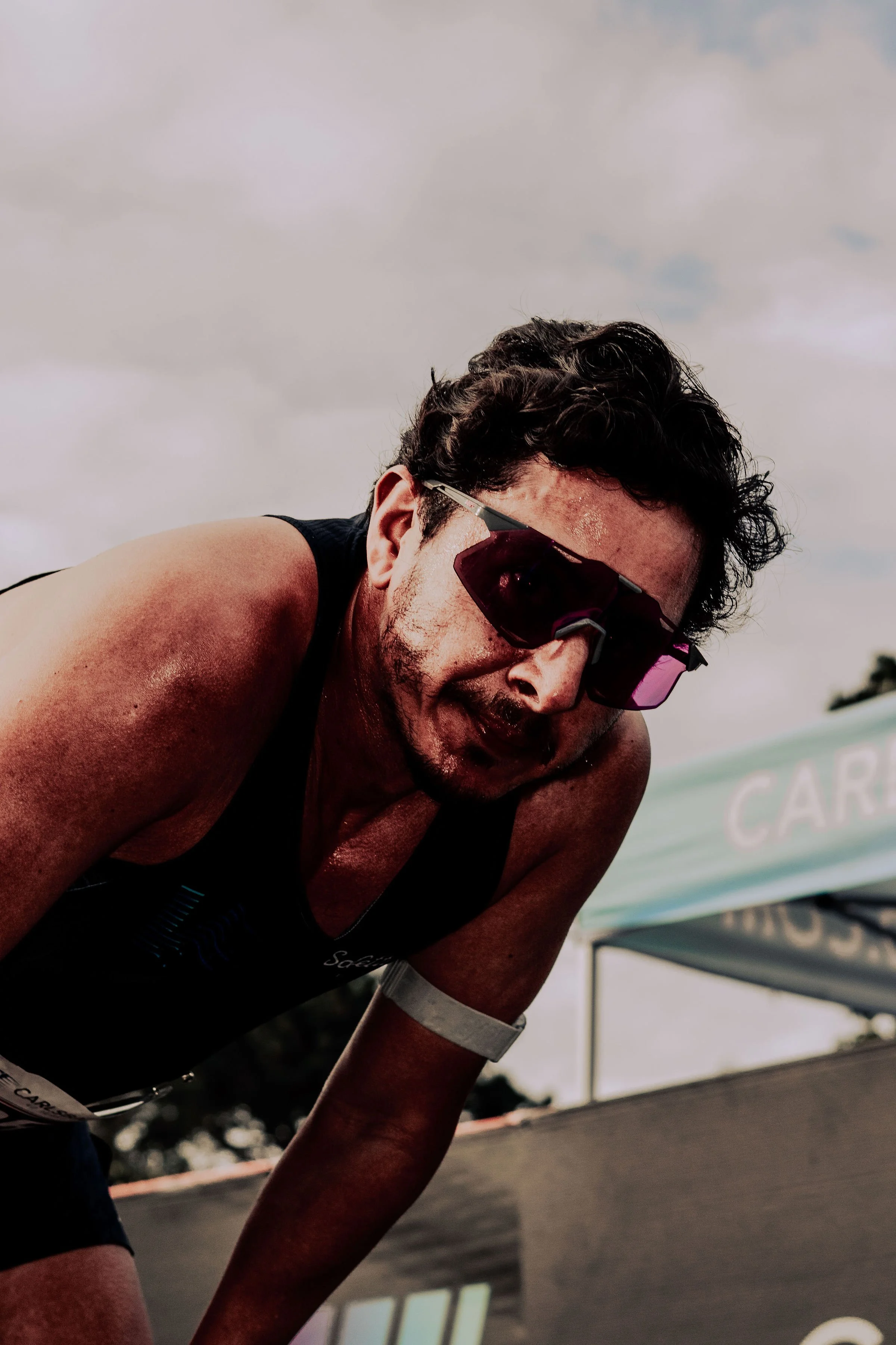 A man wearing sunglasses and a black athletic shirt, leaning forward with a focused expression, during an outdoor sports event.