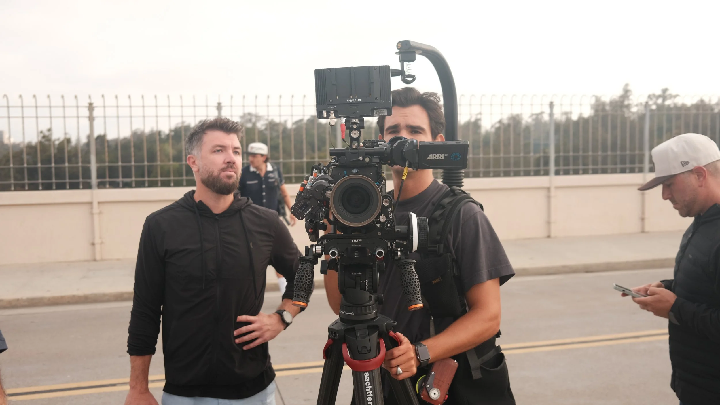 People working on a film set with a professional camera on a tripod, outdoors near a fence and road.