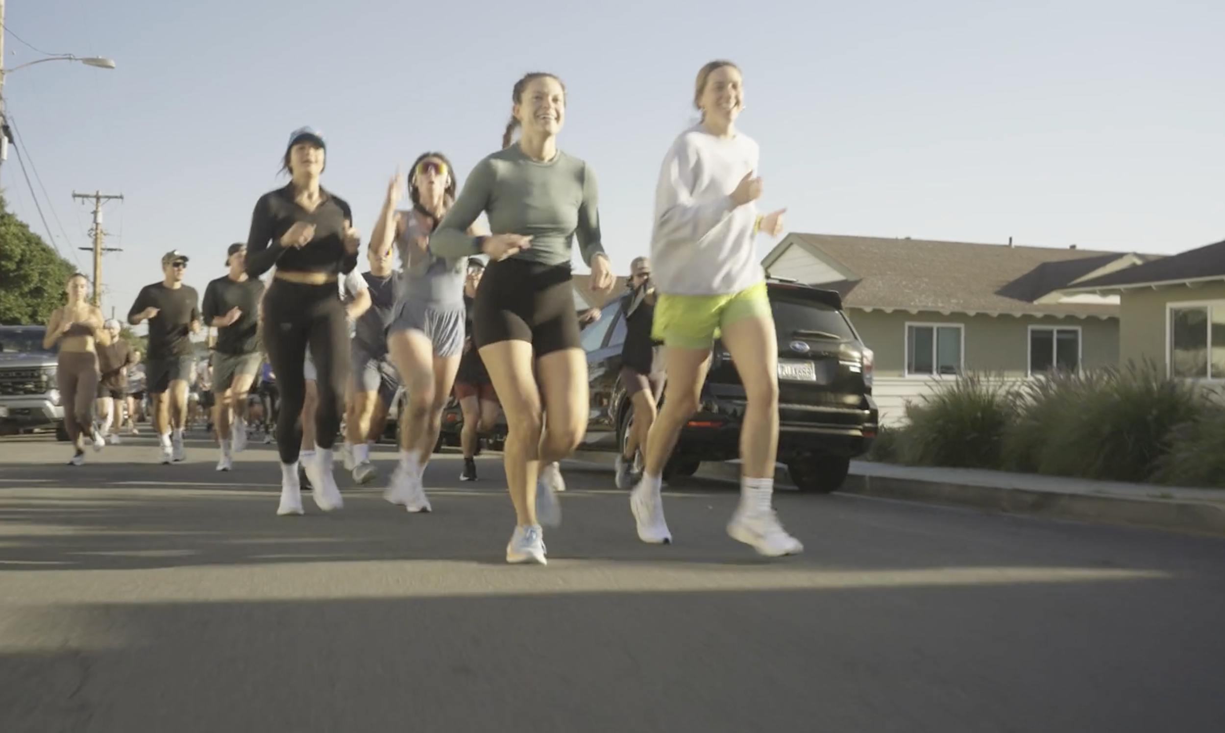 A group of women jogging on a residential street during daytime. They are wearing athletic clothing and smiling, with houses and parked cars visible in the background.