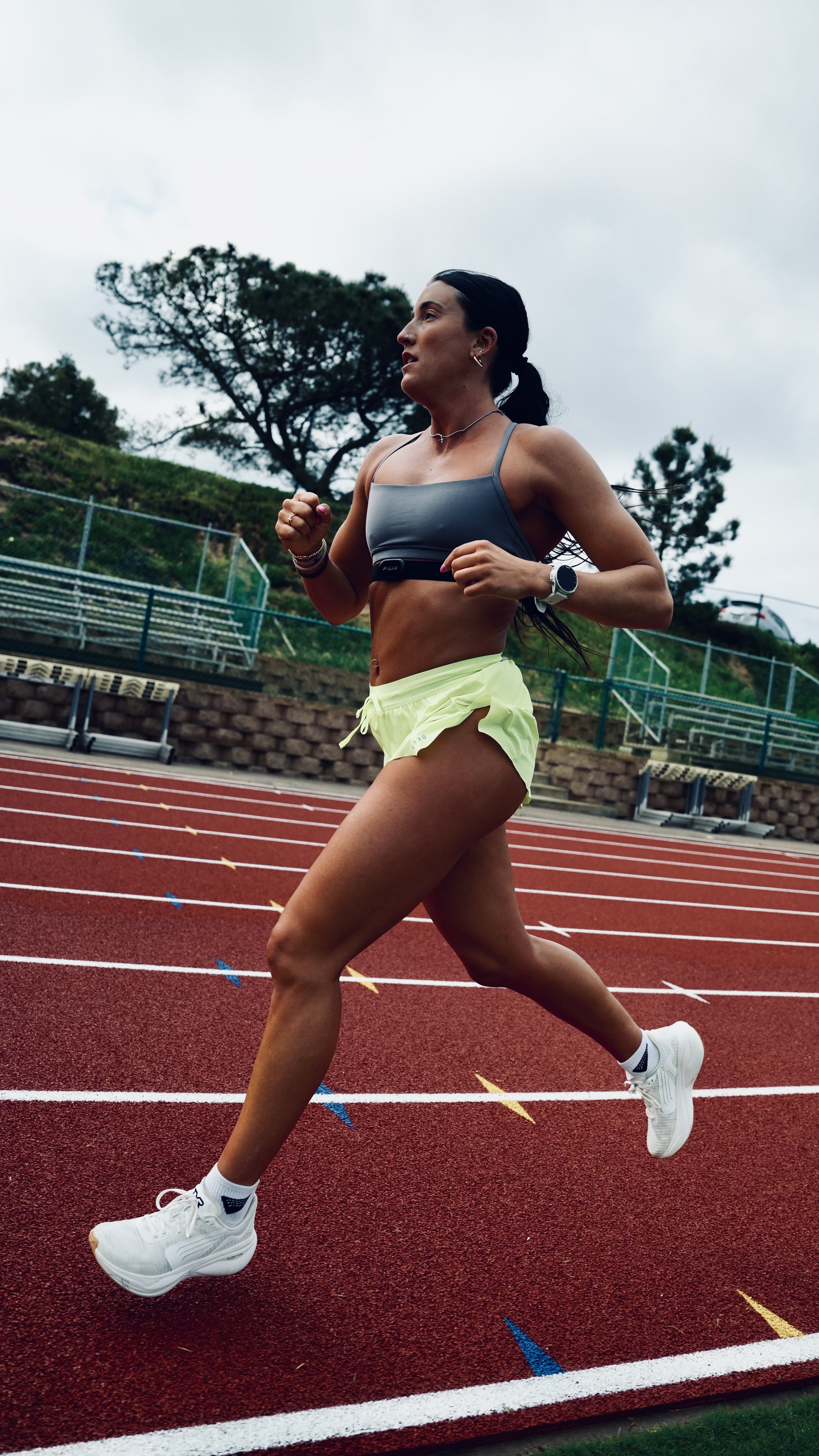 A woman running on a red outdoor track, wearing a gray sports bra, yellow shorts, white running shoes, and a smartwatch, with trees and cloudy sky in the background.
