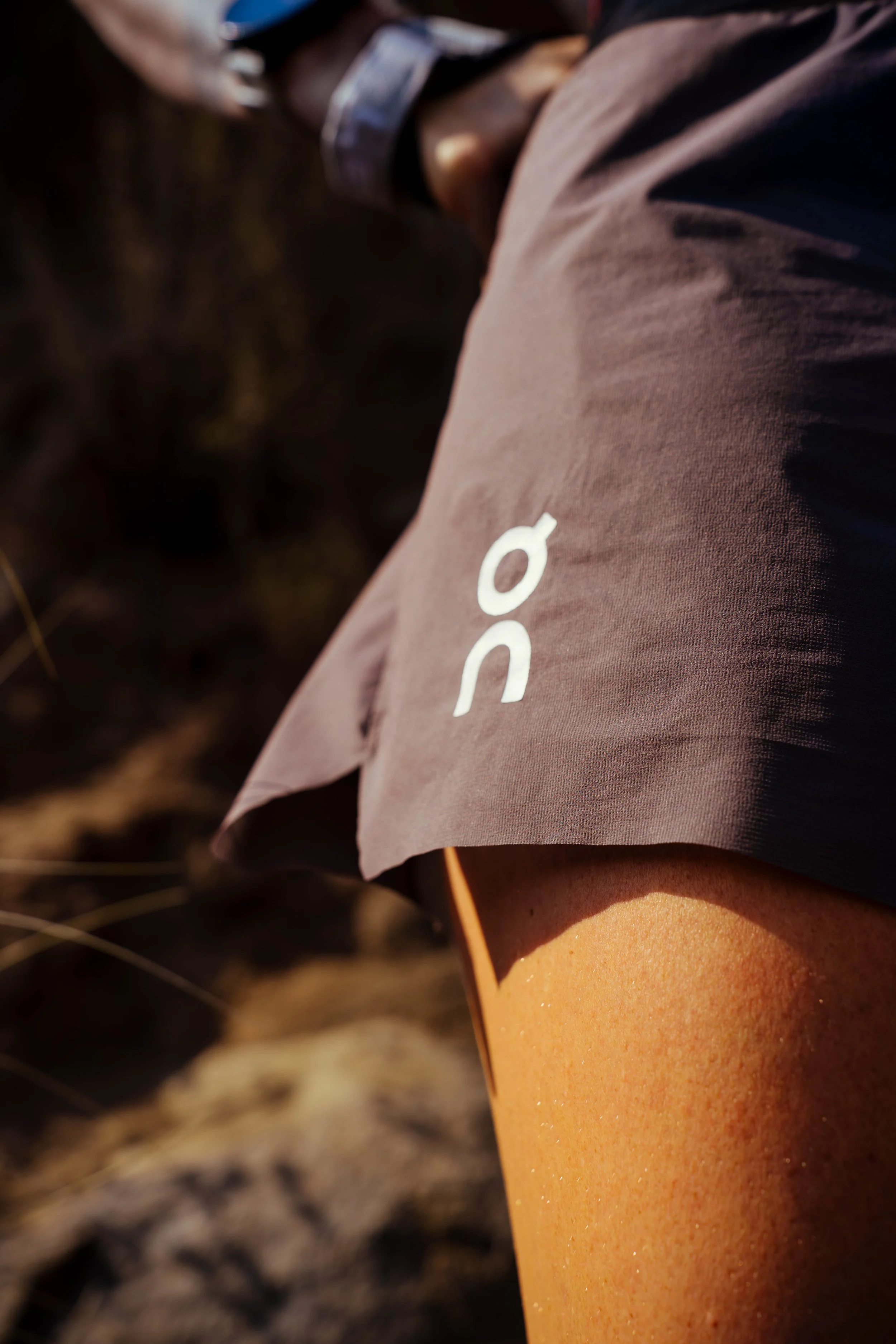 Close-up of a person's leg wearing a brown shorts and a gray wristwatch, with a gray shirt, standing on a dirt ground with dried grass.