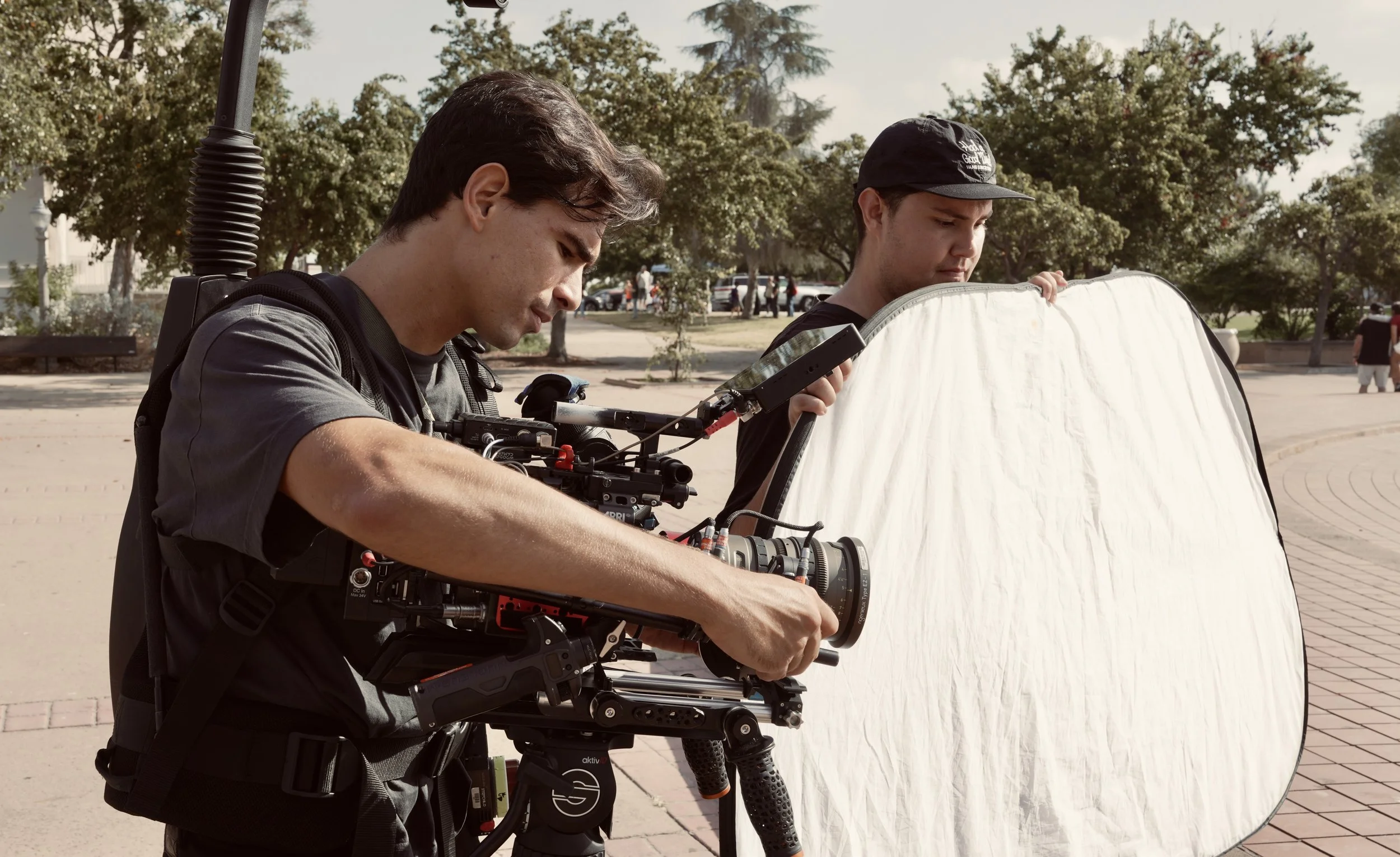 Two young men are working on filming equipment outdoors on a sunny day. One is adjusting a camera mounted on a stabilizer, the other is holding a reflective diffuser to regulate lighting. There are trees and a few people in the background.