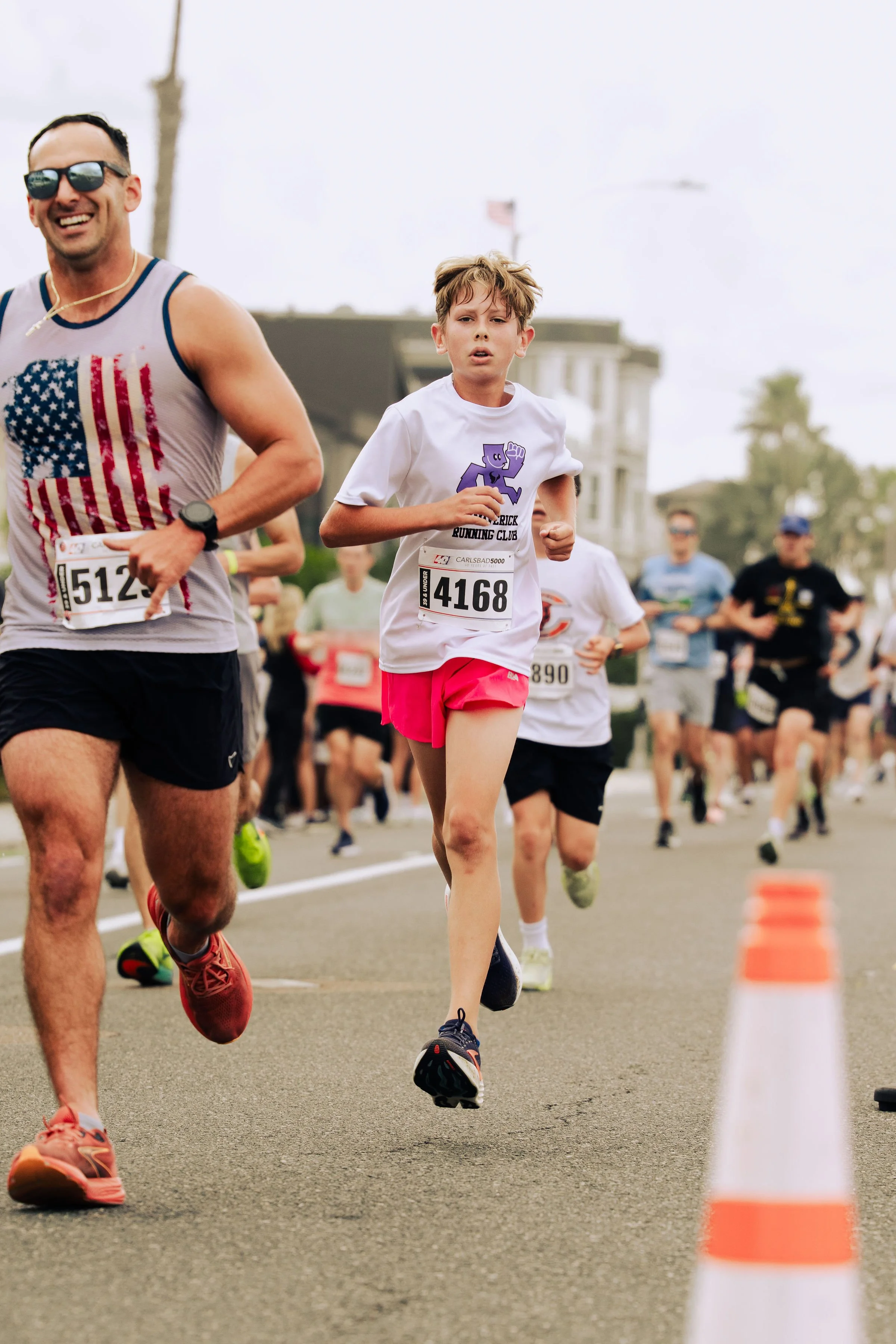 People participating in a road race or marathon, running on a city street with a traffic cone in the foreground, overcast sky, and buildings in the background.