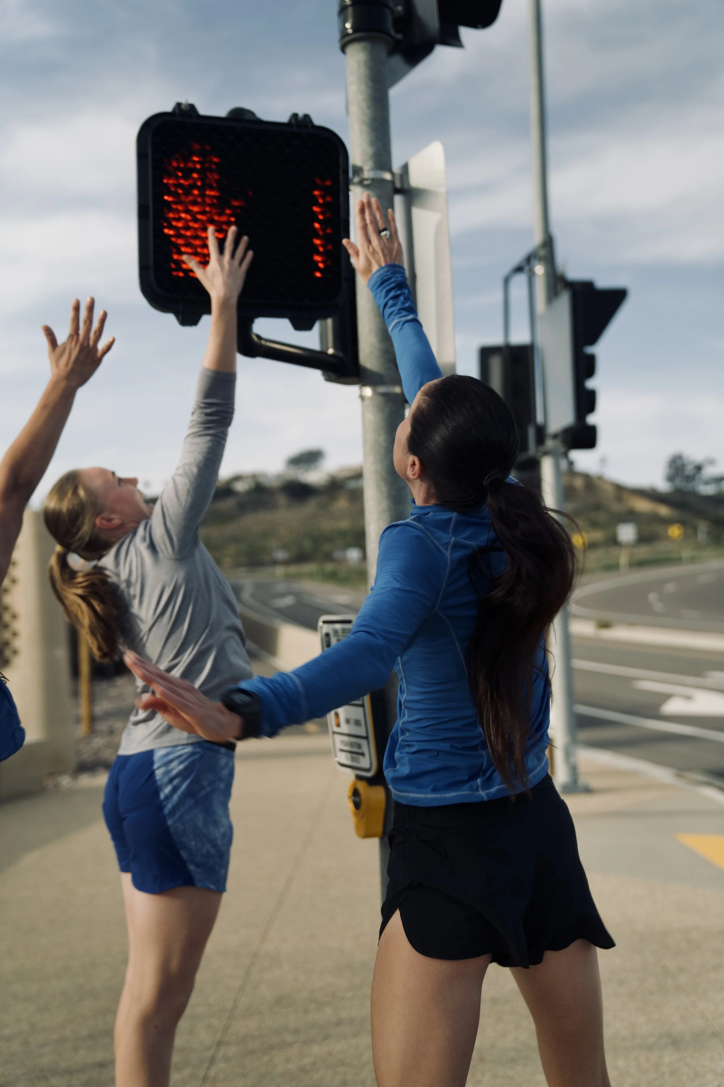 Two women stretch their legs and arms near a red traffic light on a street, with a cloudy sky above.