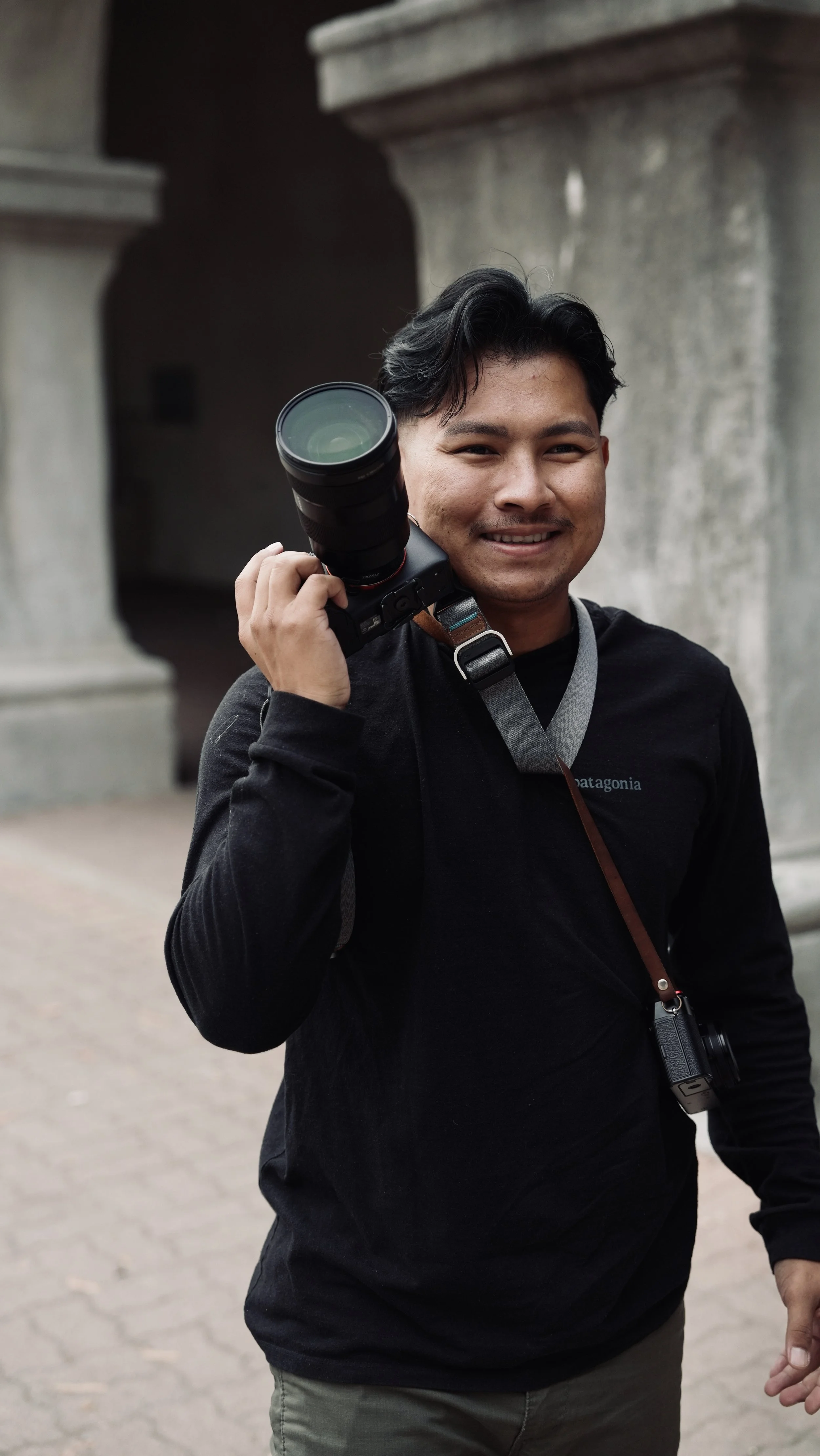 A smiling man holding a camera with a telephoto lens over his shoulder outdoors near stone columns.