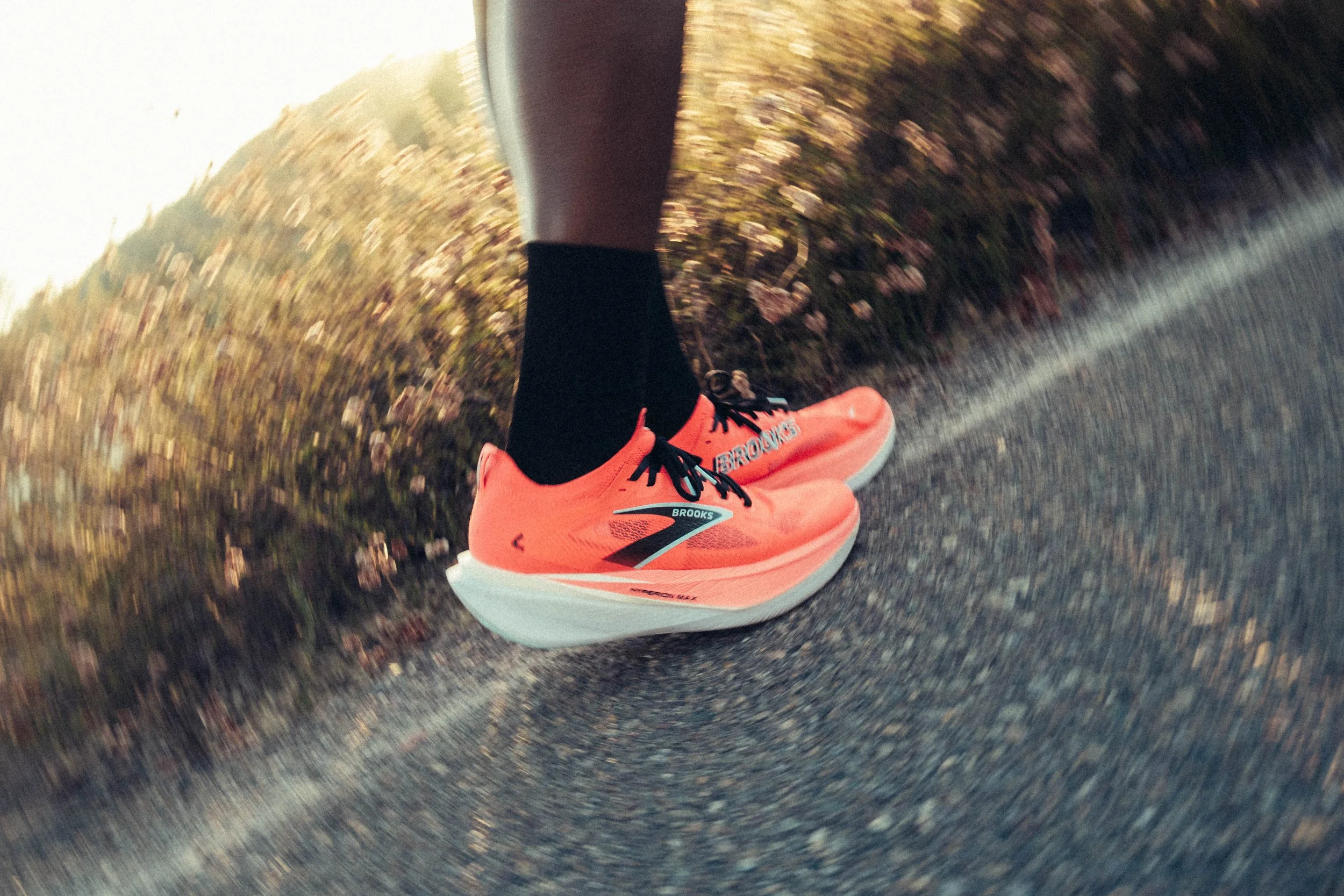 Close-up of a person's foot wearing a bright orange Brooks running shoe, black sock, and gray leggings, running on a paved road with blurred vegetation on the side.