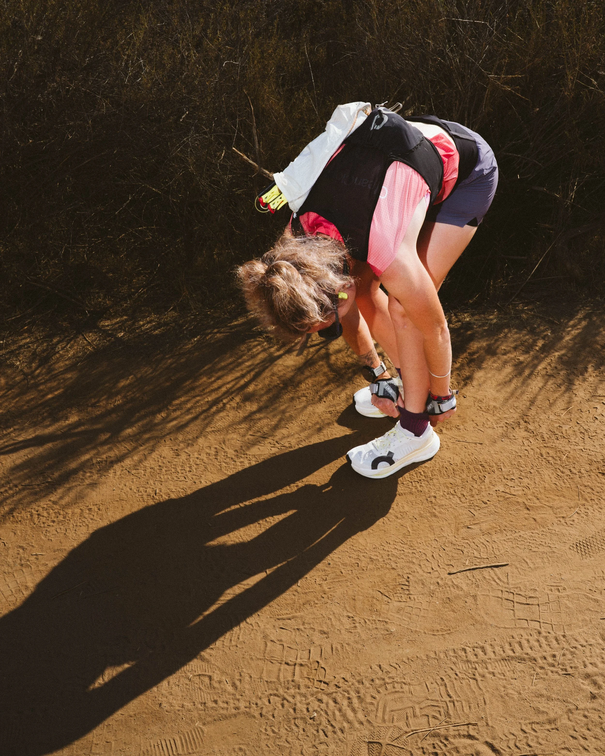A woman tying her shoelaces on a dirt trail, with her shadow cast on the ground and a background of dry bushes.