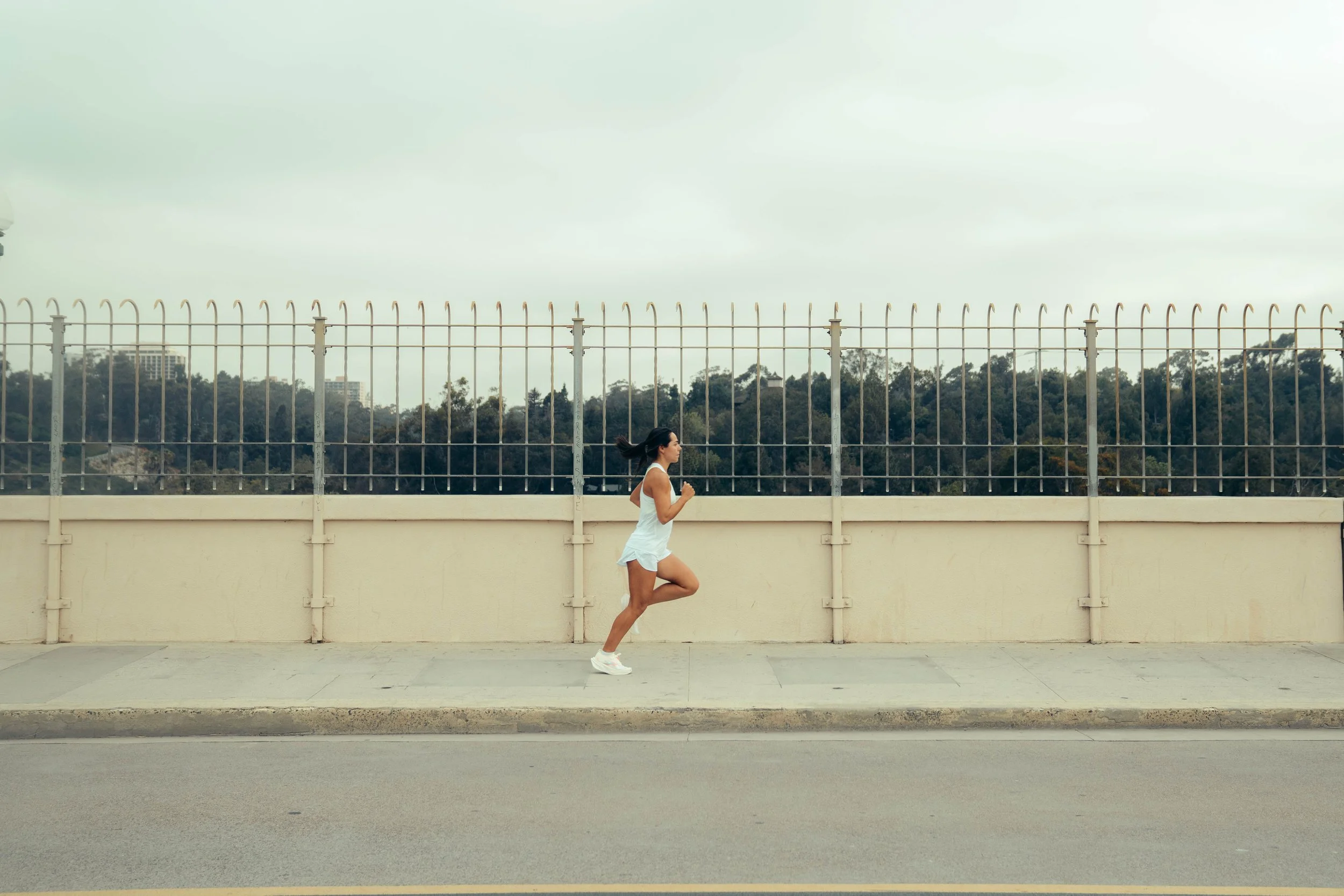 A woman jogging on a sidewalk in front of a beige wall and metal fence with trees and buildings in the background.