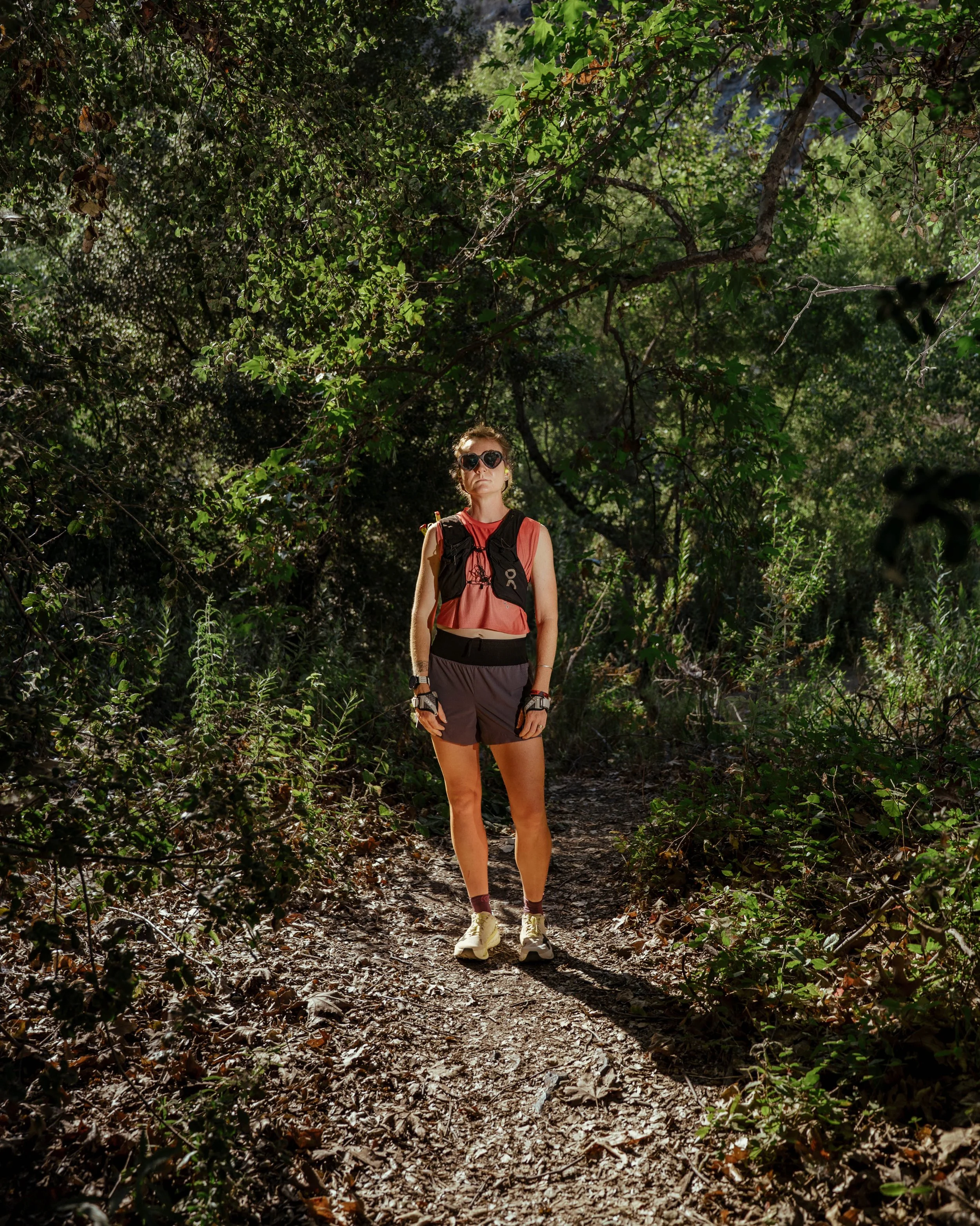 A woman with sunglasses, a pink sleeveless top, black shorts, and a backpack walking on a dirt trail through a dense, leafy forest.