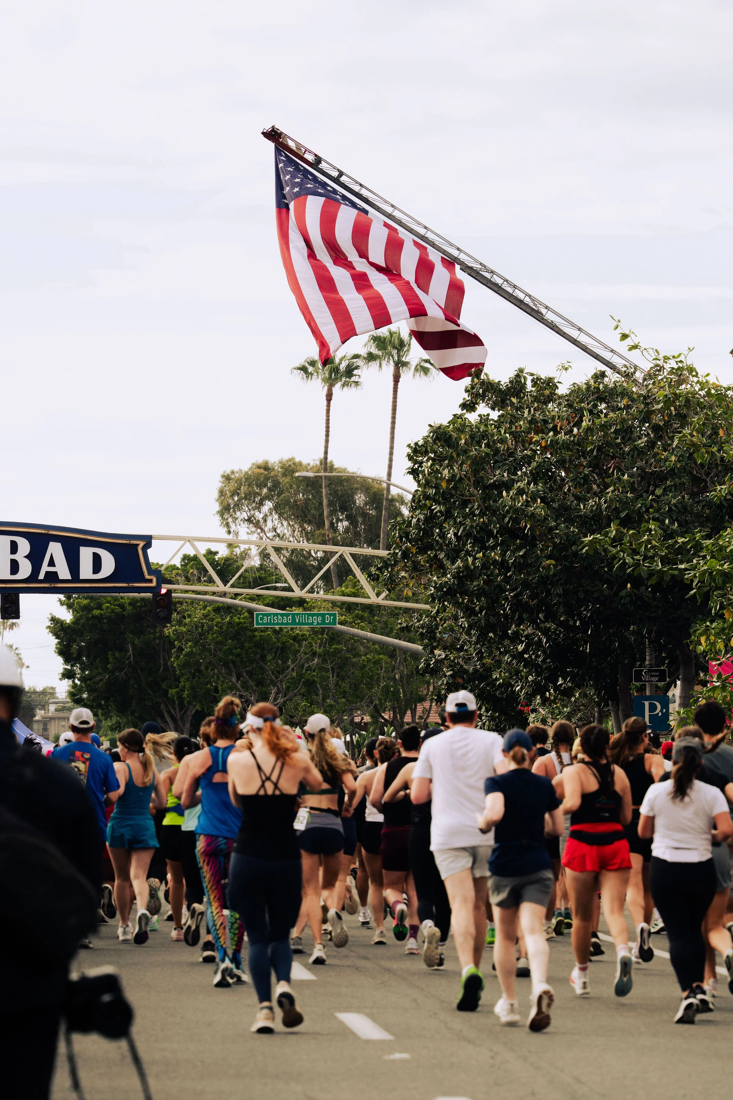 Crowd of people running outside during a race or marathon, with an American flag hanging from a fire truck and palm trees in the background.
