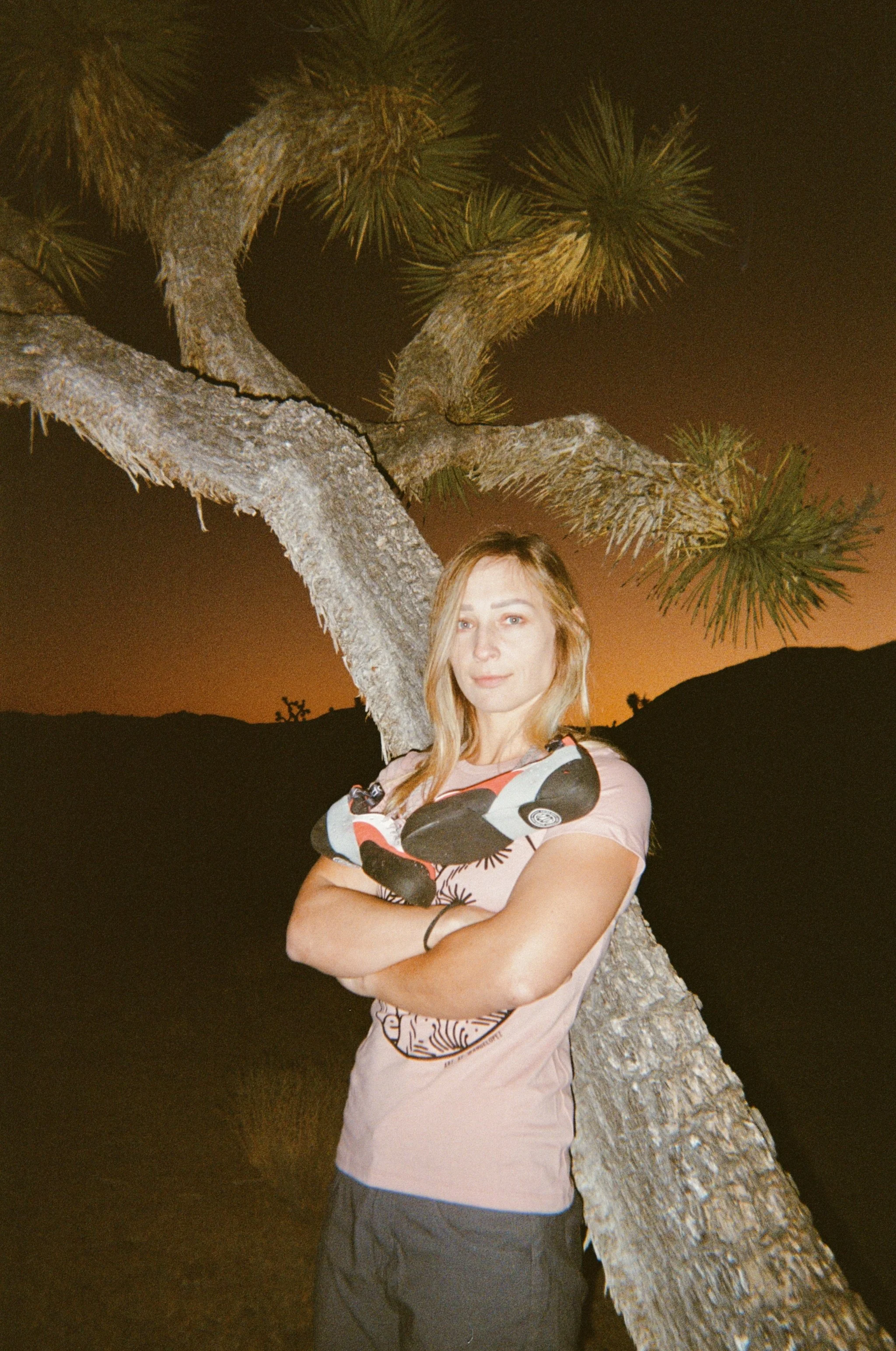 A woman with long blonde hair and a pink T-shirt leaning against a tree at sunset or dusk, with a dark landscape and sky in the background.