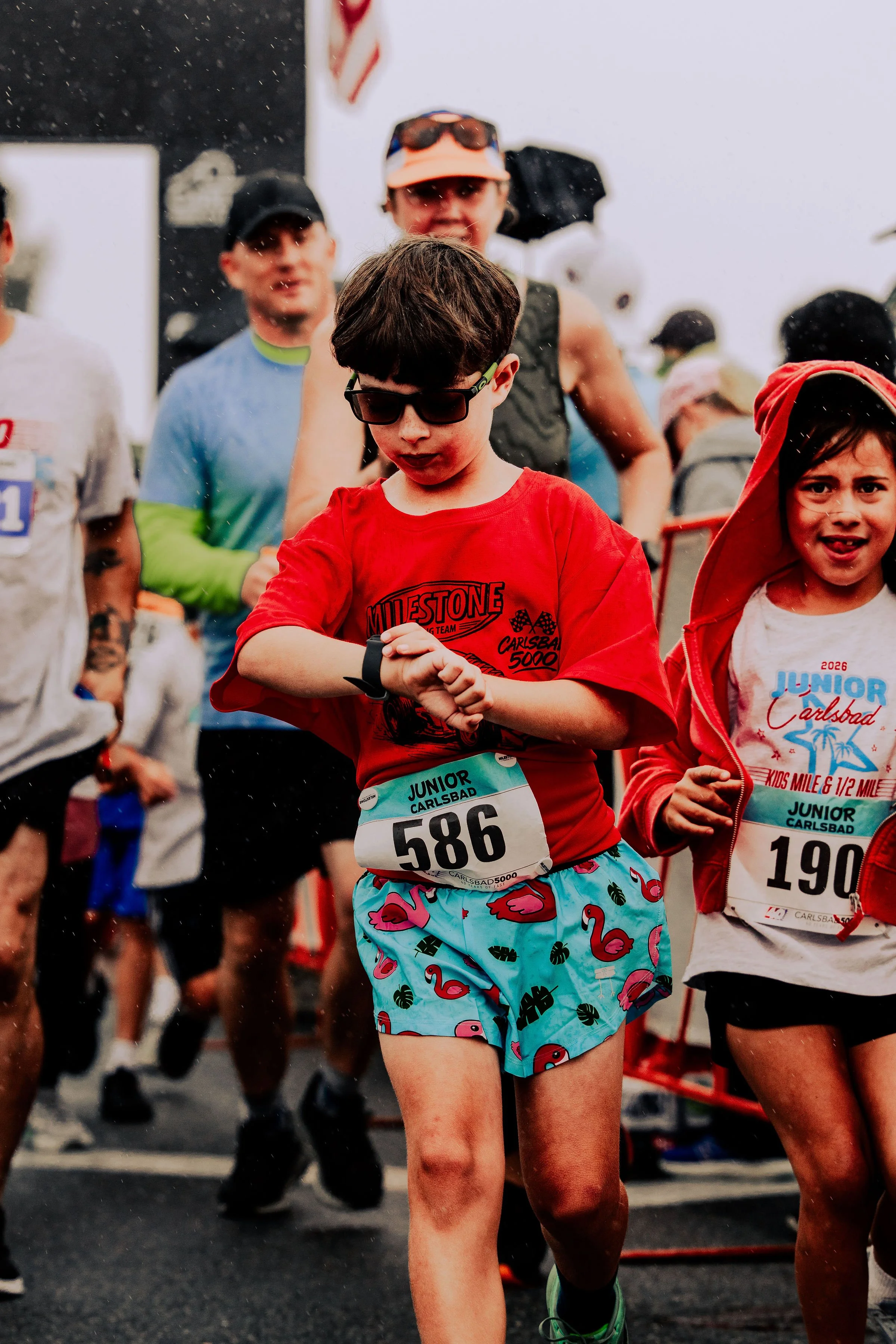 Participants in a running event, with children and adults, wearing race bibs, casual athletic clothes, and sunglasses, on a wet, overcast day.