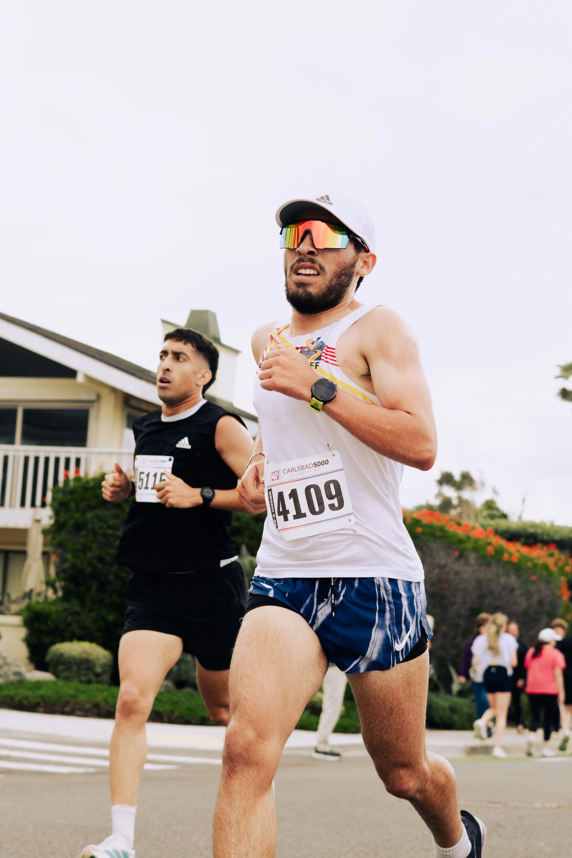 Two male runners participating in a road race, wearing athletic clothing and race bibs, with one in the foreground and the other slightly behind, with a background of residential houses and landscaping.