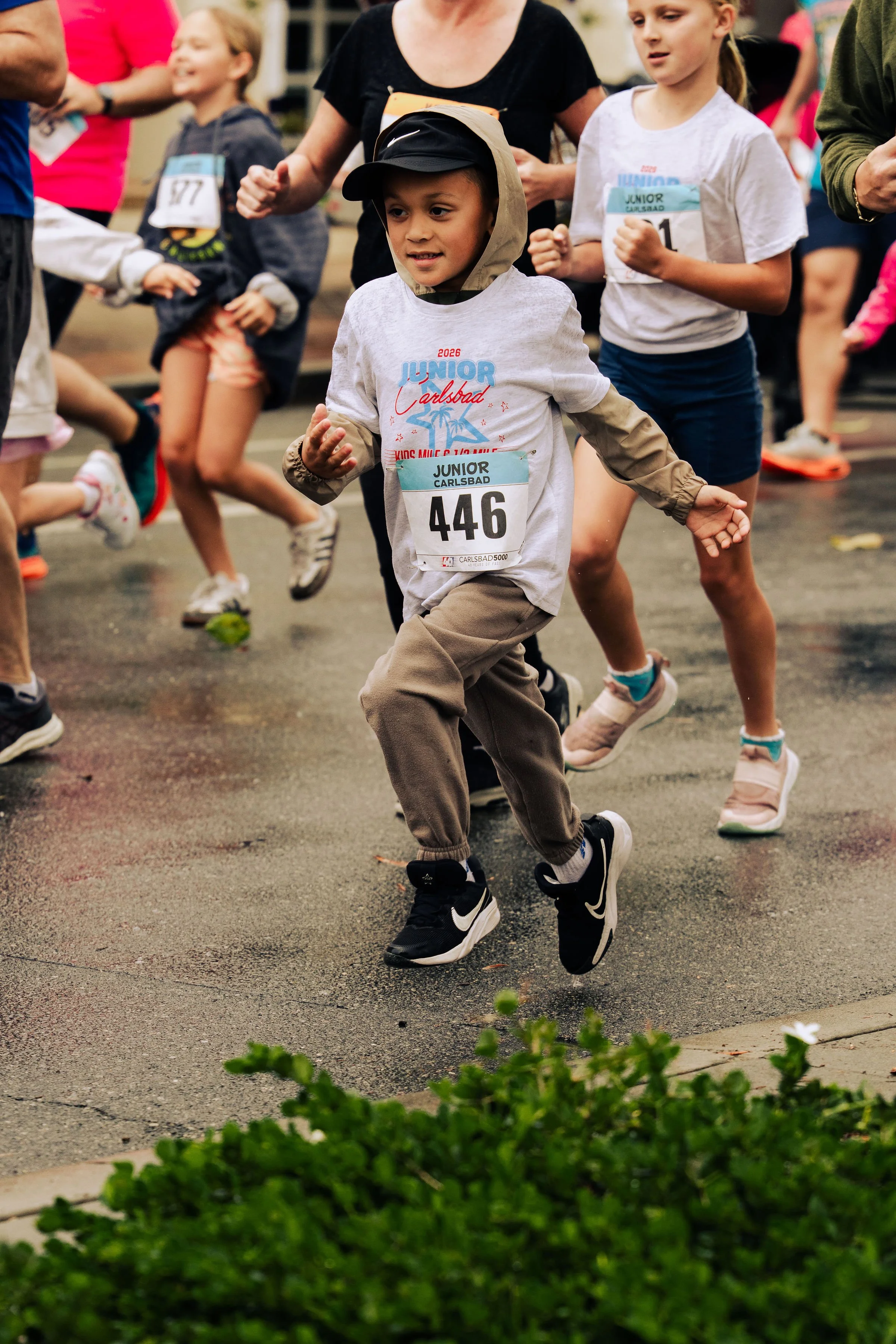 Young boy wearing a hoodie and running shoes participating in a children's race, surrounded by other kids and adults, with a focus on him in the middle of the race. The background shows a festive environment, possibly a park or community event.