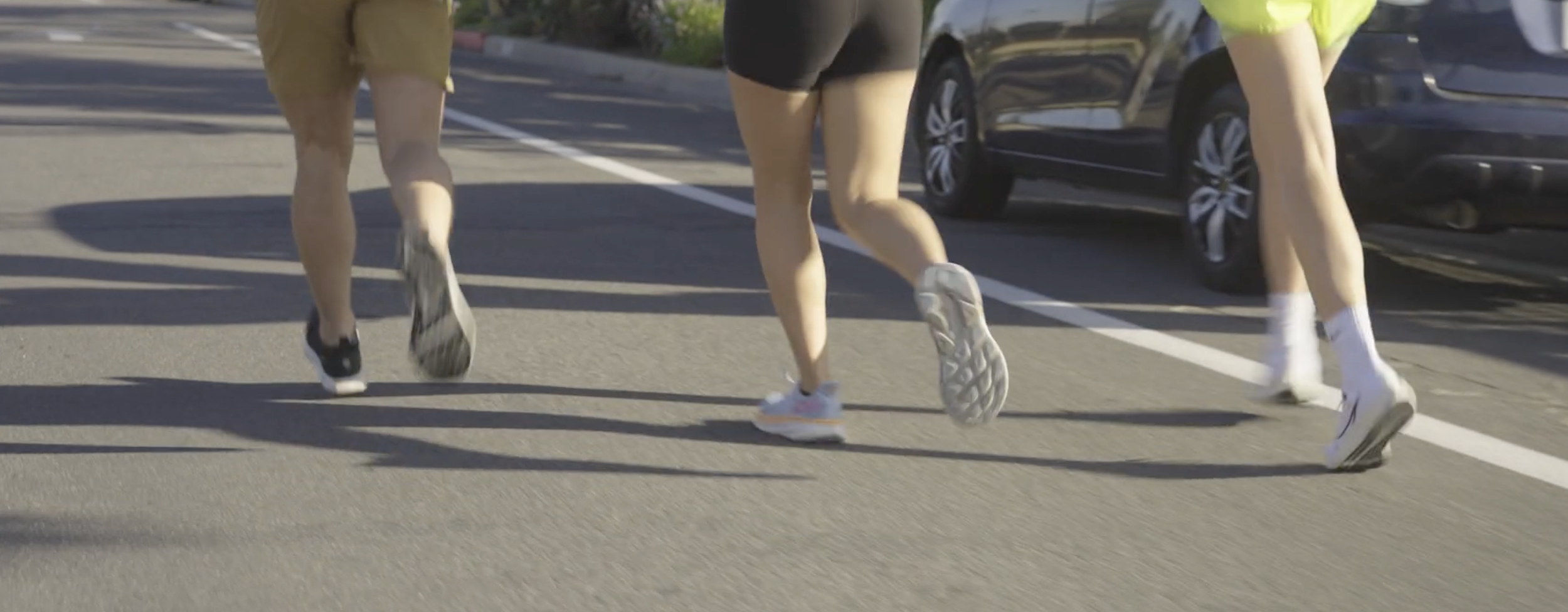 Three people running on a parking lot, wearing athletic shoes and shorts, with parked cars in the background.