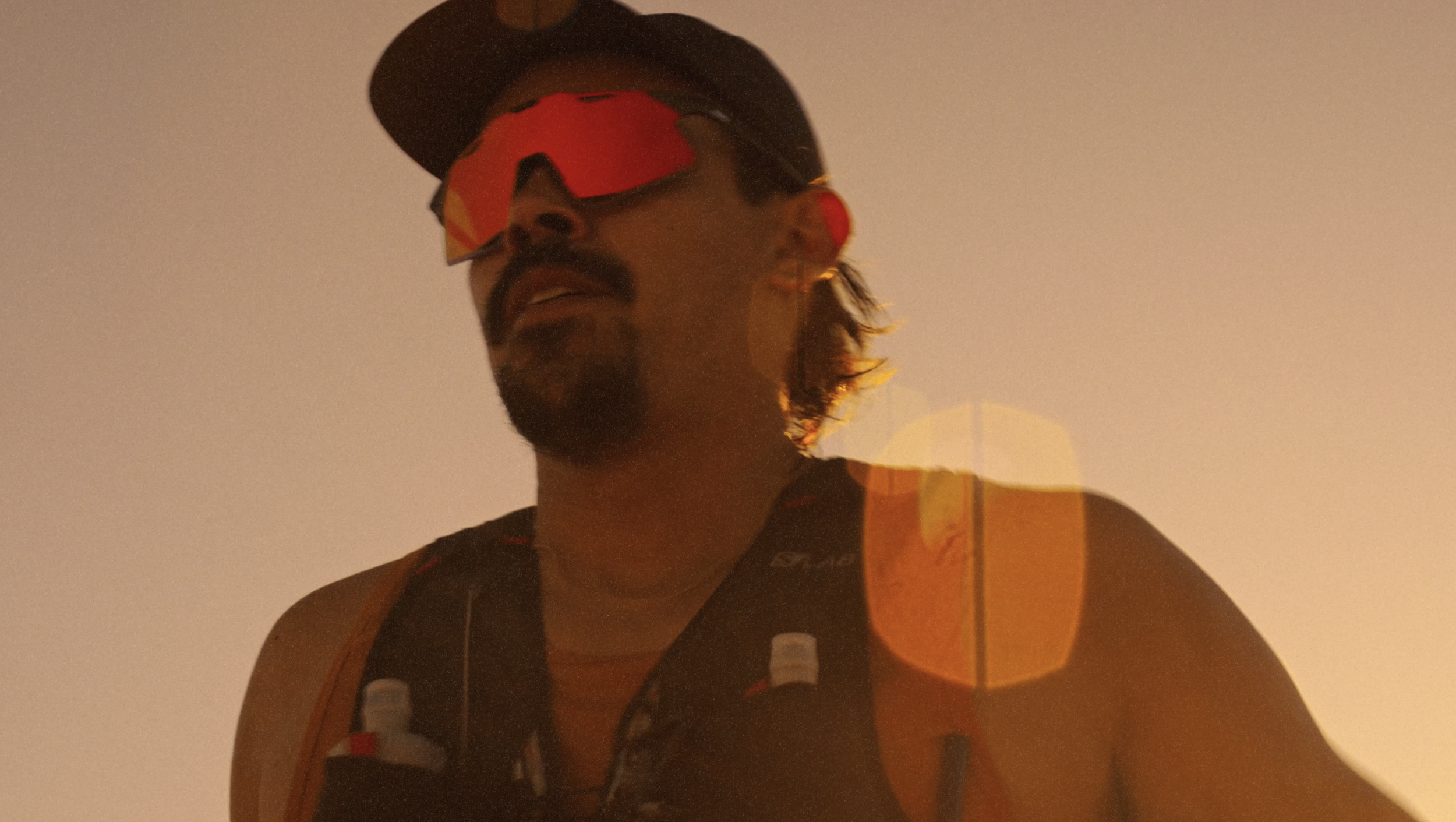 A person wearing a black cap, sunglasses, and a sleeveless shirt, standing against a plain background with warm lighting.