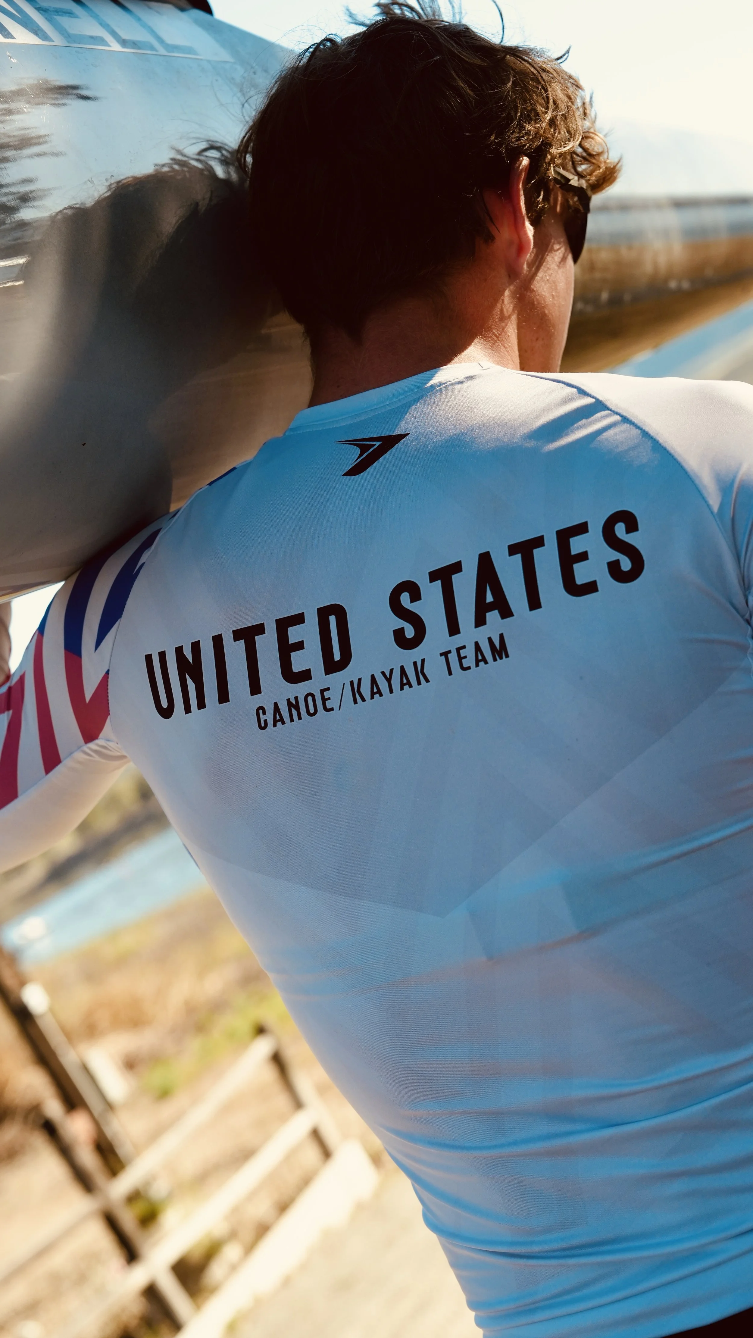 Back view of a person wearing a white USA kayak team shirt, holding a kayak, outdoors near water.