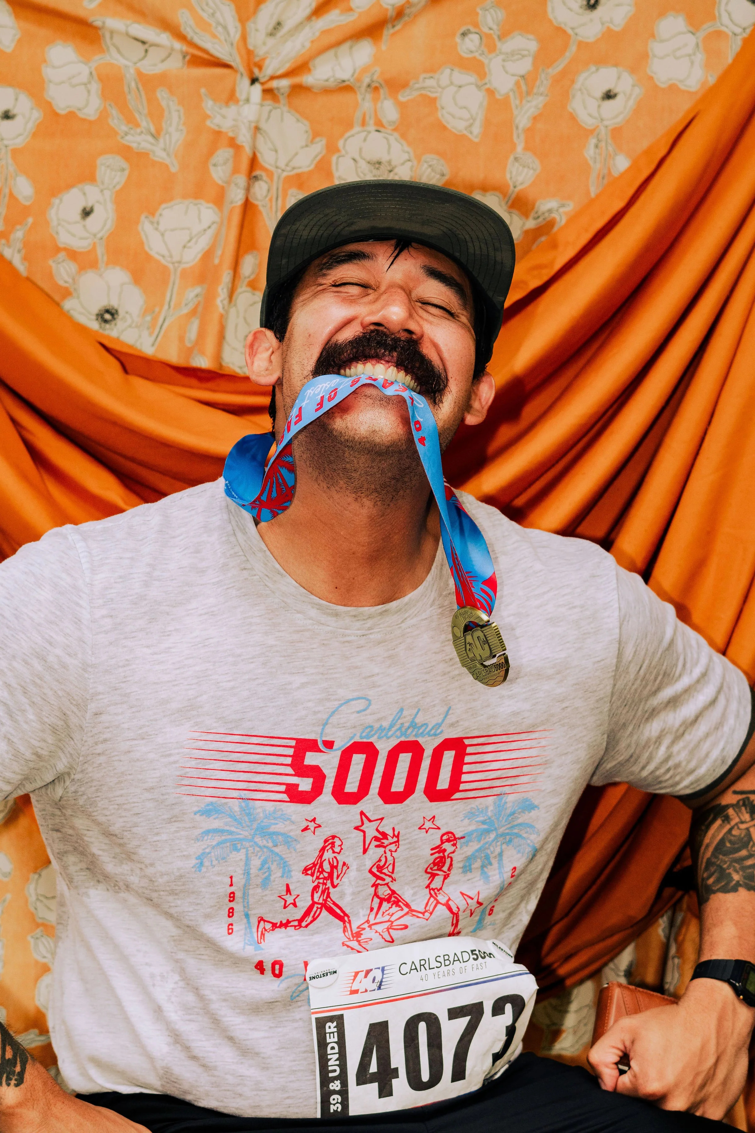Man smiling with a medal in his mouth, wearing a gray T-shirt and a cap, with a floral orange backdrop.