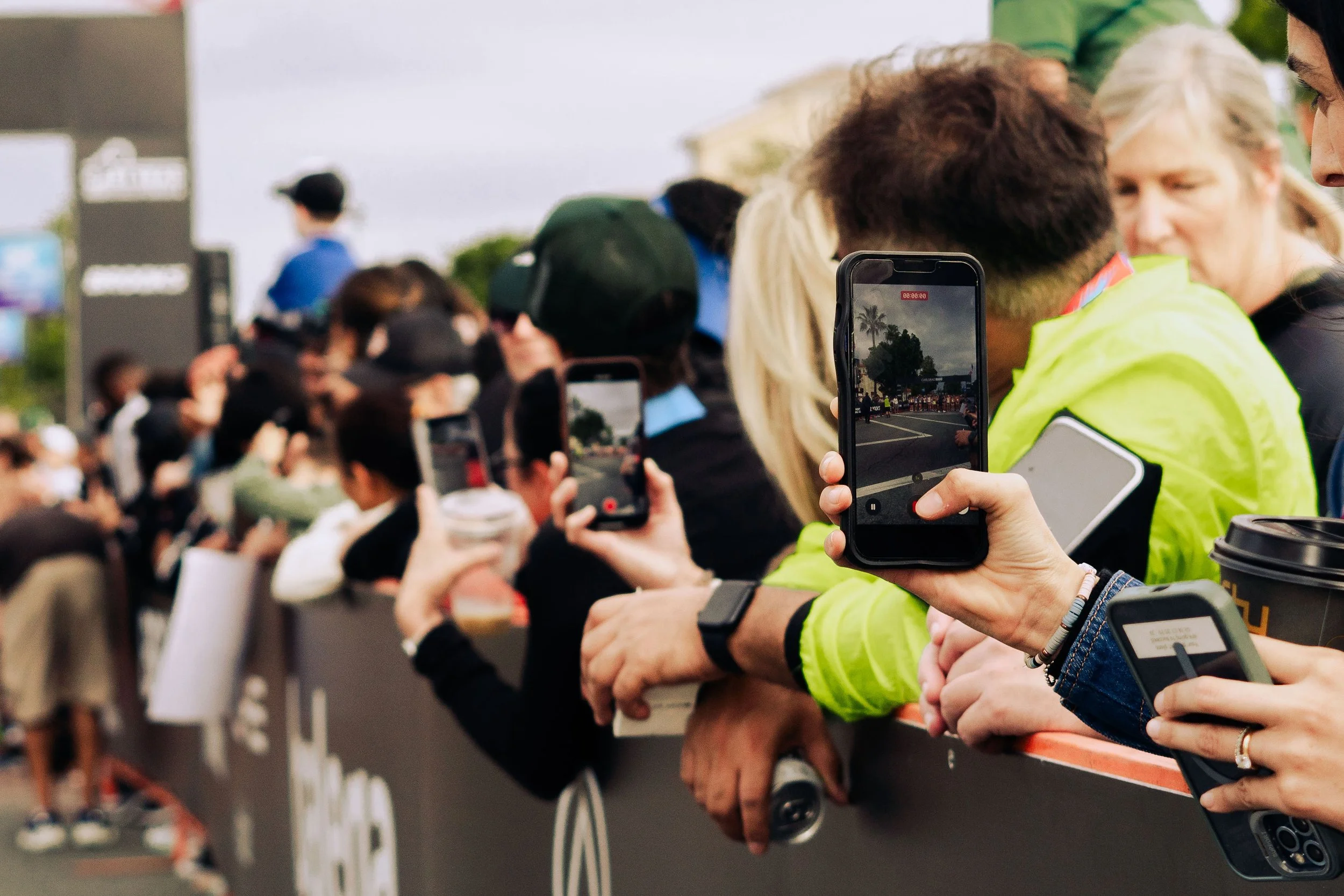 Crowd of people at an outdoor event, many holding up phones, some wearing high-visibility jackets, with a barricade separating them from the event area.