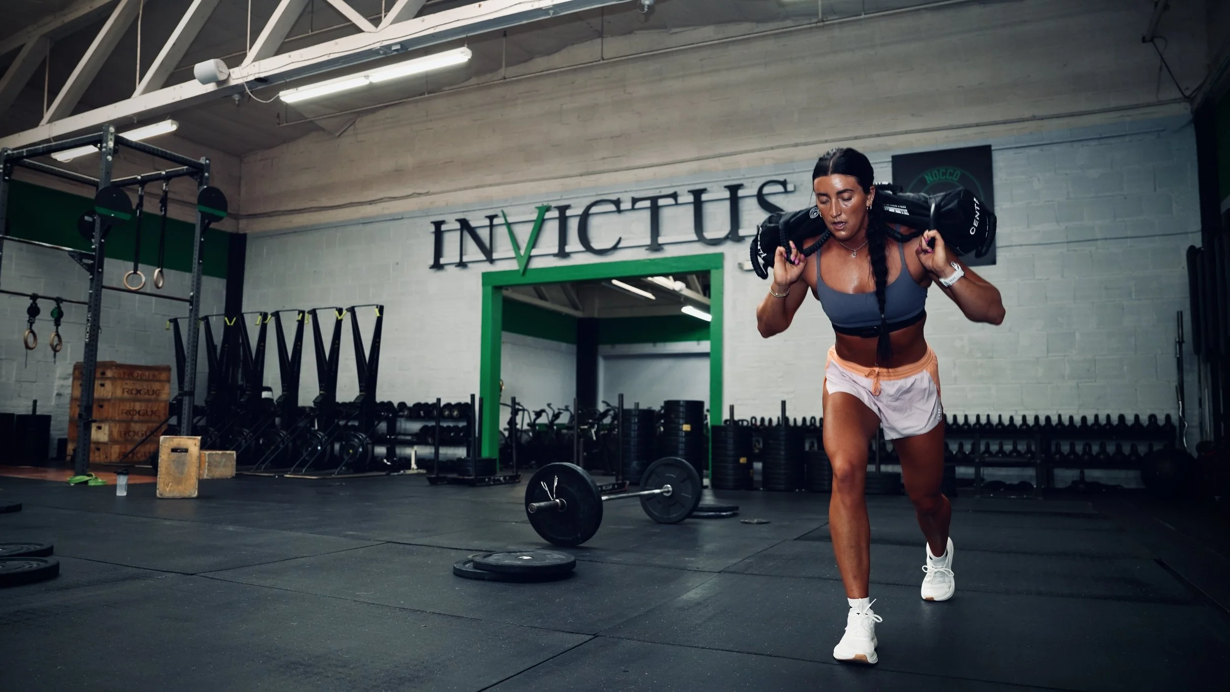 A woman in workout clothes lifting a heavy sandbag on her shoulders inside a gym, with fitness equipment and weights in the background.