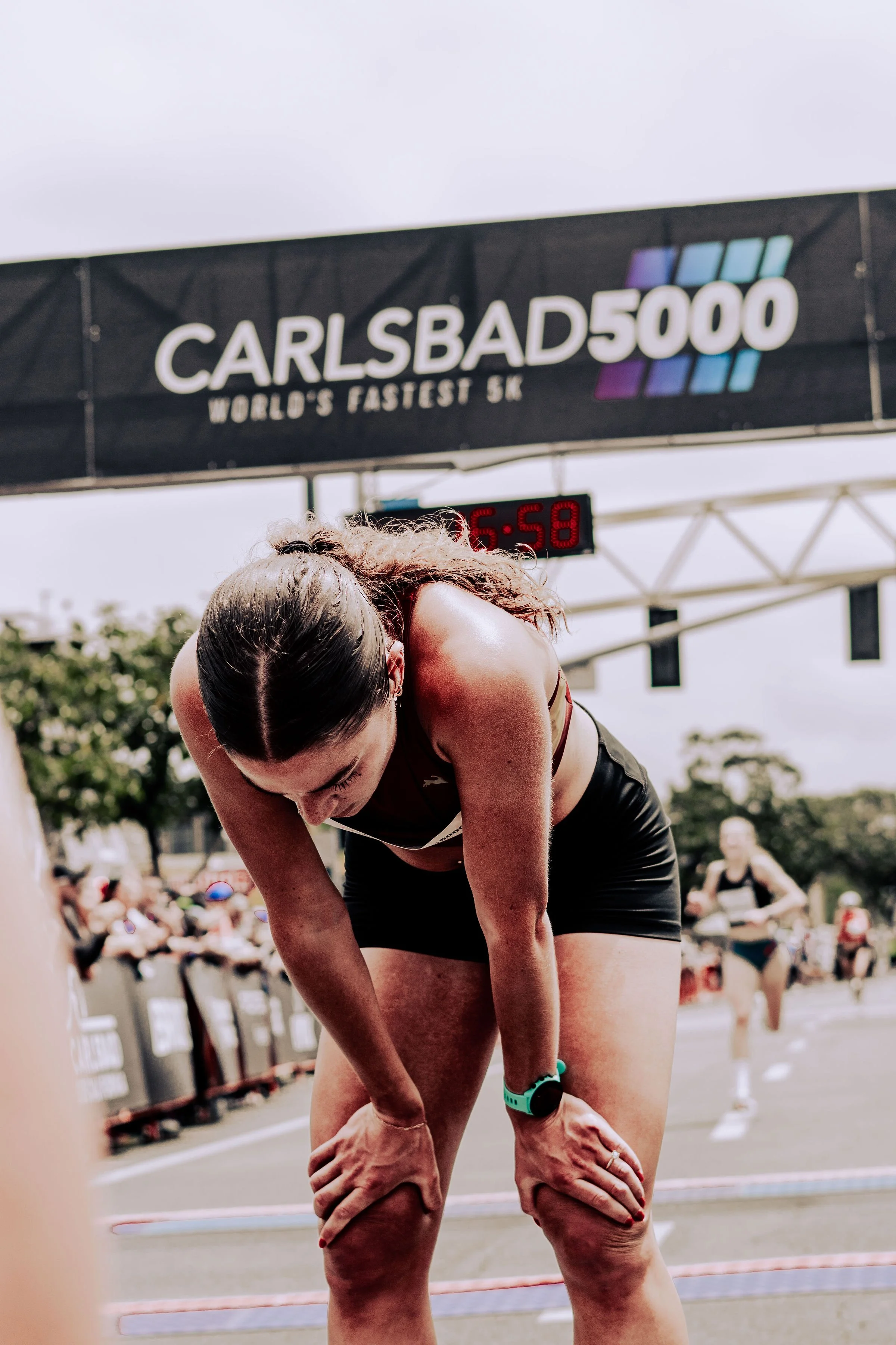 A female athlete is bent over, resting her hands on her knees, during a race or sporting event. She is wearing a sports watch and a black sports bra. In the background, there are spectators and a digital timer showing 6:58. A large overhead sign read