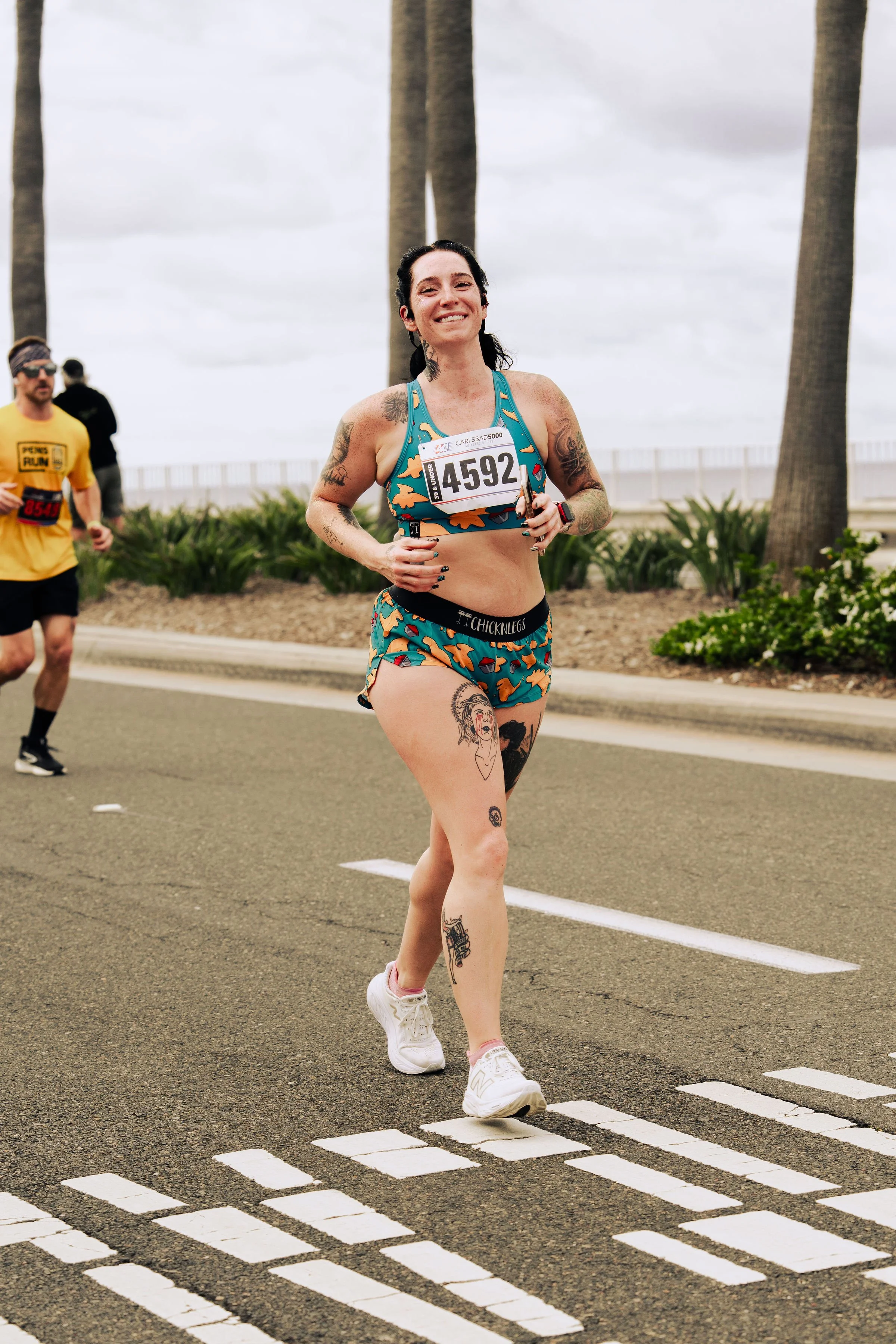 Female runner smiling while participating in a race, wearing a colorful sports outfit and running on a street with palm trees in the background.
