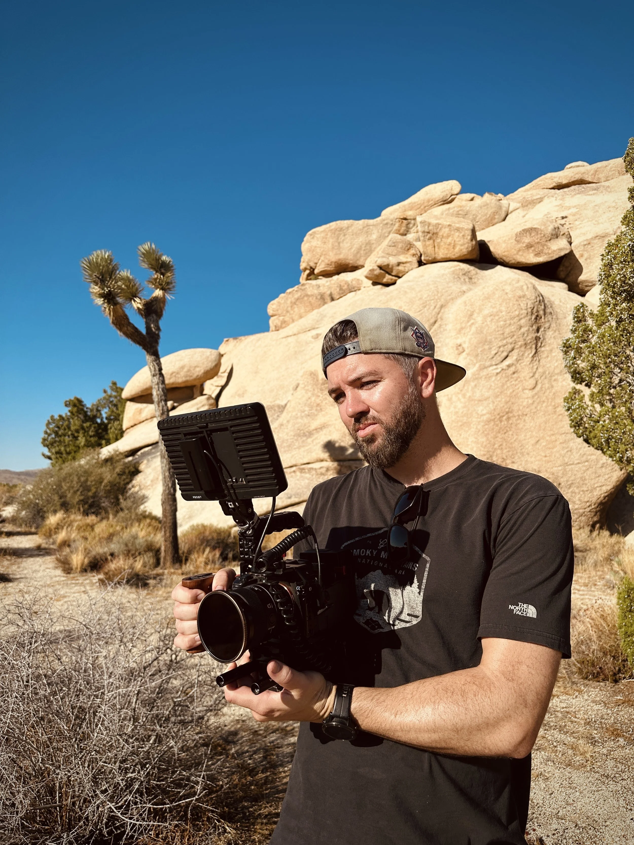 A man wearing a baseball cap and a t-shirt stands outdoors in a desert landscape with large rocks, a Joshua tree, and dry bushes, holding a camera stabilizer and looking at a monitor.