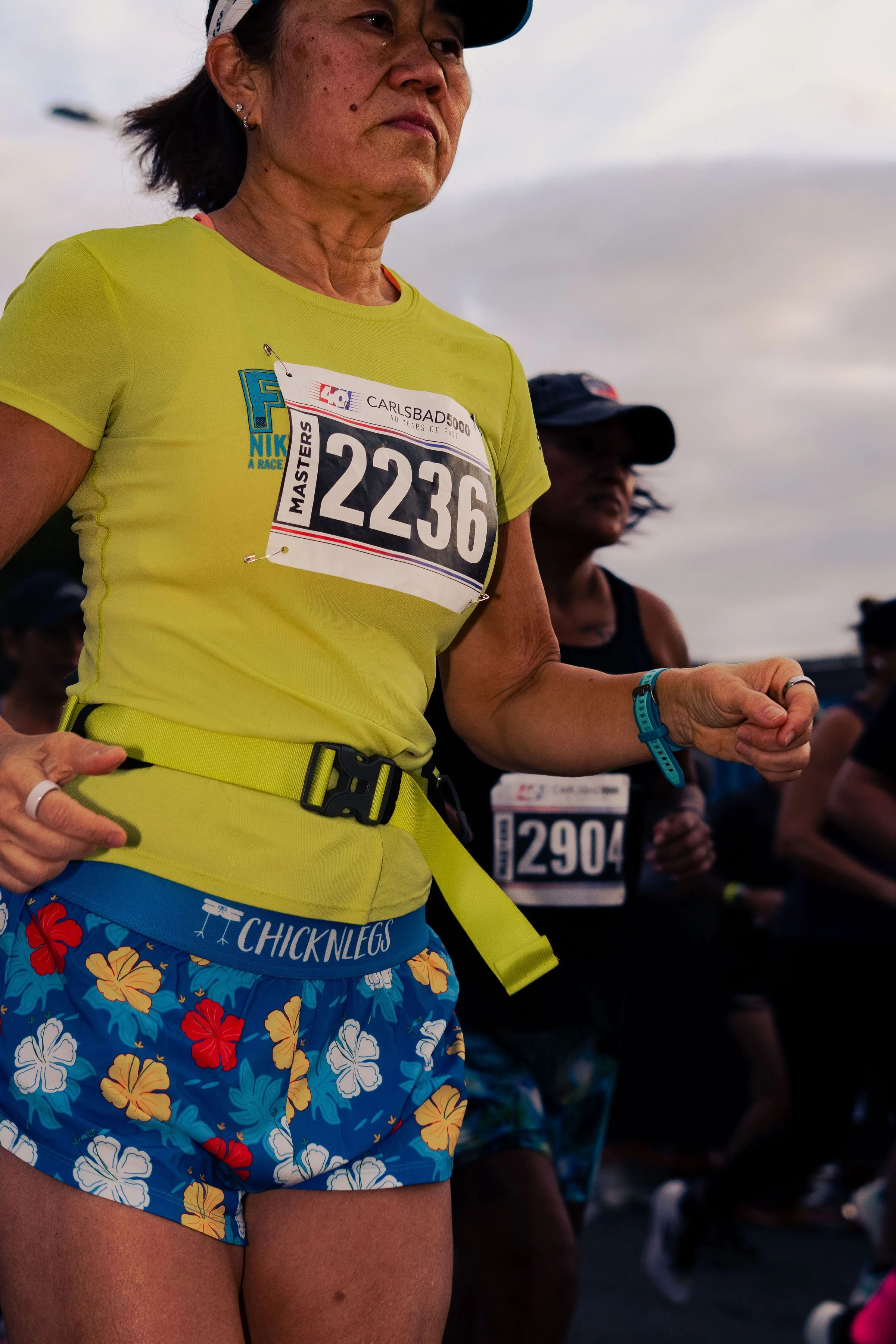 A woman wearing a yellow shirt with a race bib number 2236, blue floral shorts, and colorful tropical-themed shorts. There are other race participants in the background.