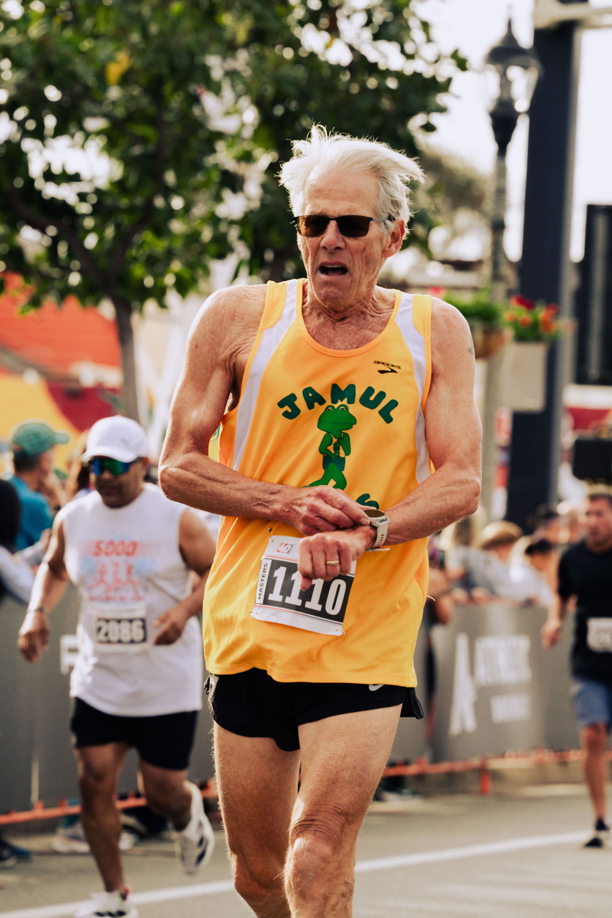 An elderly man with white hair and sunglasses participating in a marathon or race, wearing a yellow singlet with a frog mascot and the word 'Jamul' on it.