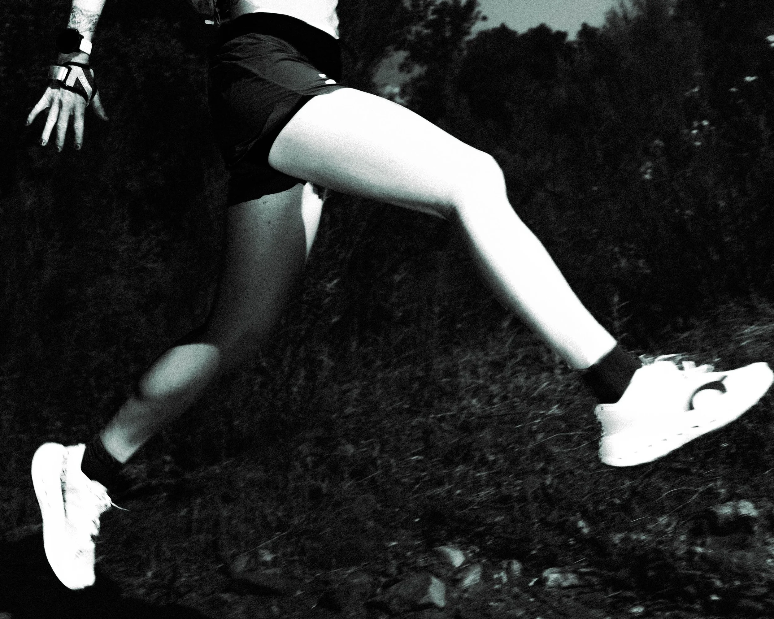 Black and white photo of a person's legs and feet lying on the grass, wearing athletic shoes and shorts, with trees in the background.