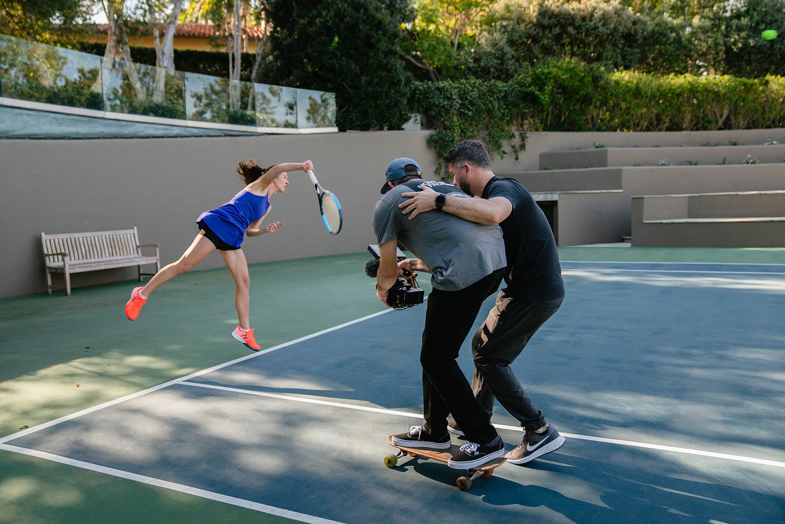 Two men filming a young woman playing tennis on a court with a skateboard, capturing her mid-air swing.