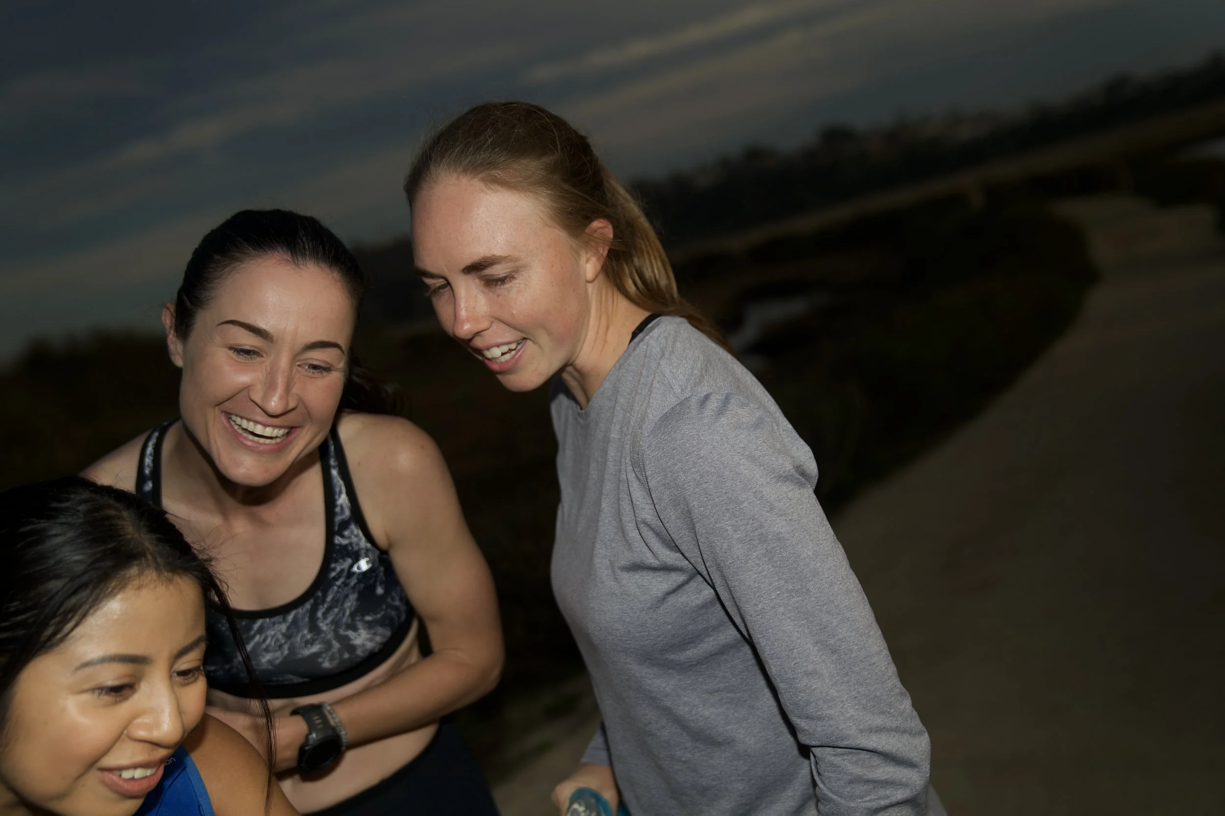 Two women and a girl smiling and looking downwards at an outdoor event during dusk.